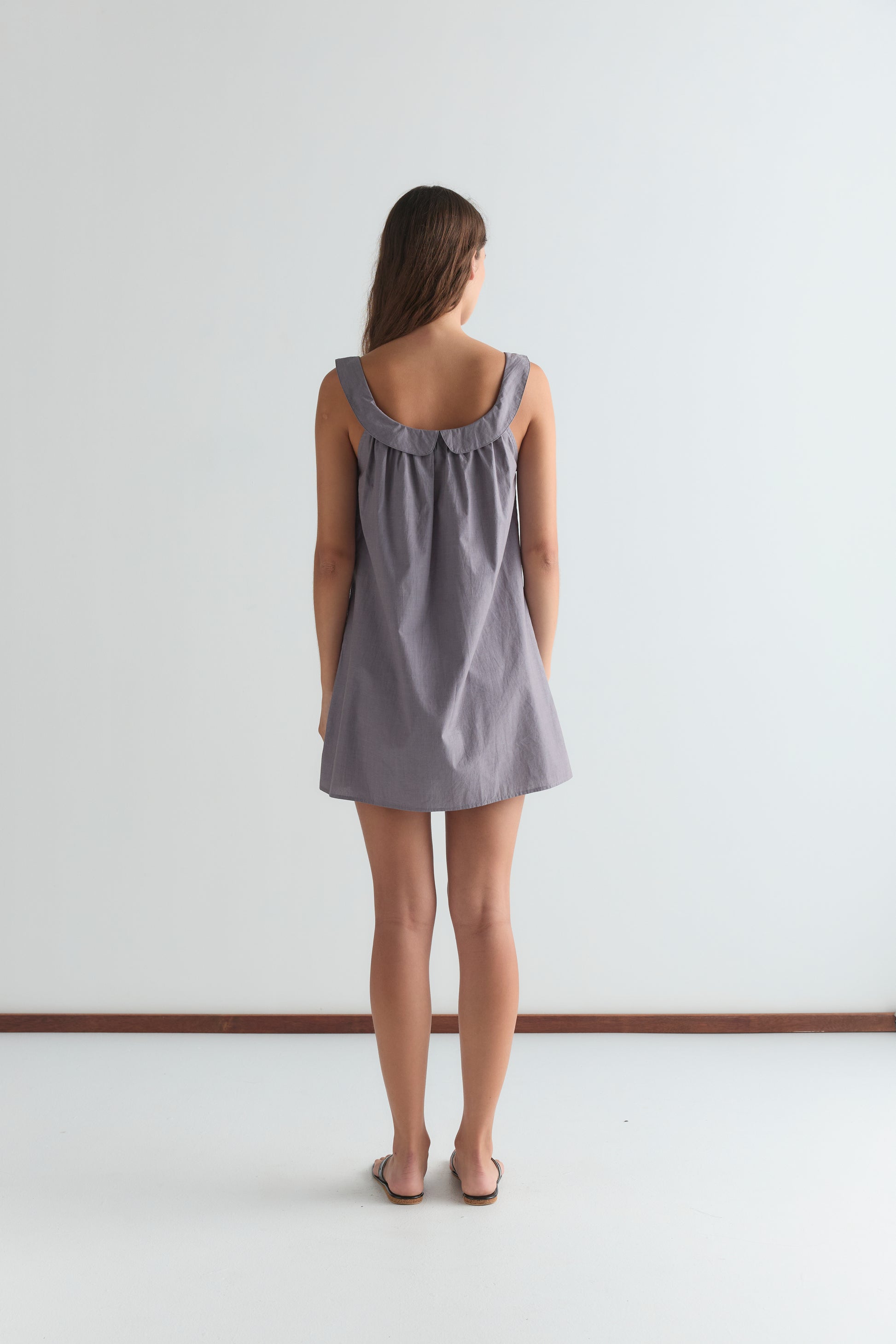 A woman faces away from the camera, wearing the Rounded Neck Dress - Currant, a loose sleeveless style with a gathered neckline, paired with brown sandals, against a plain white background.