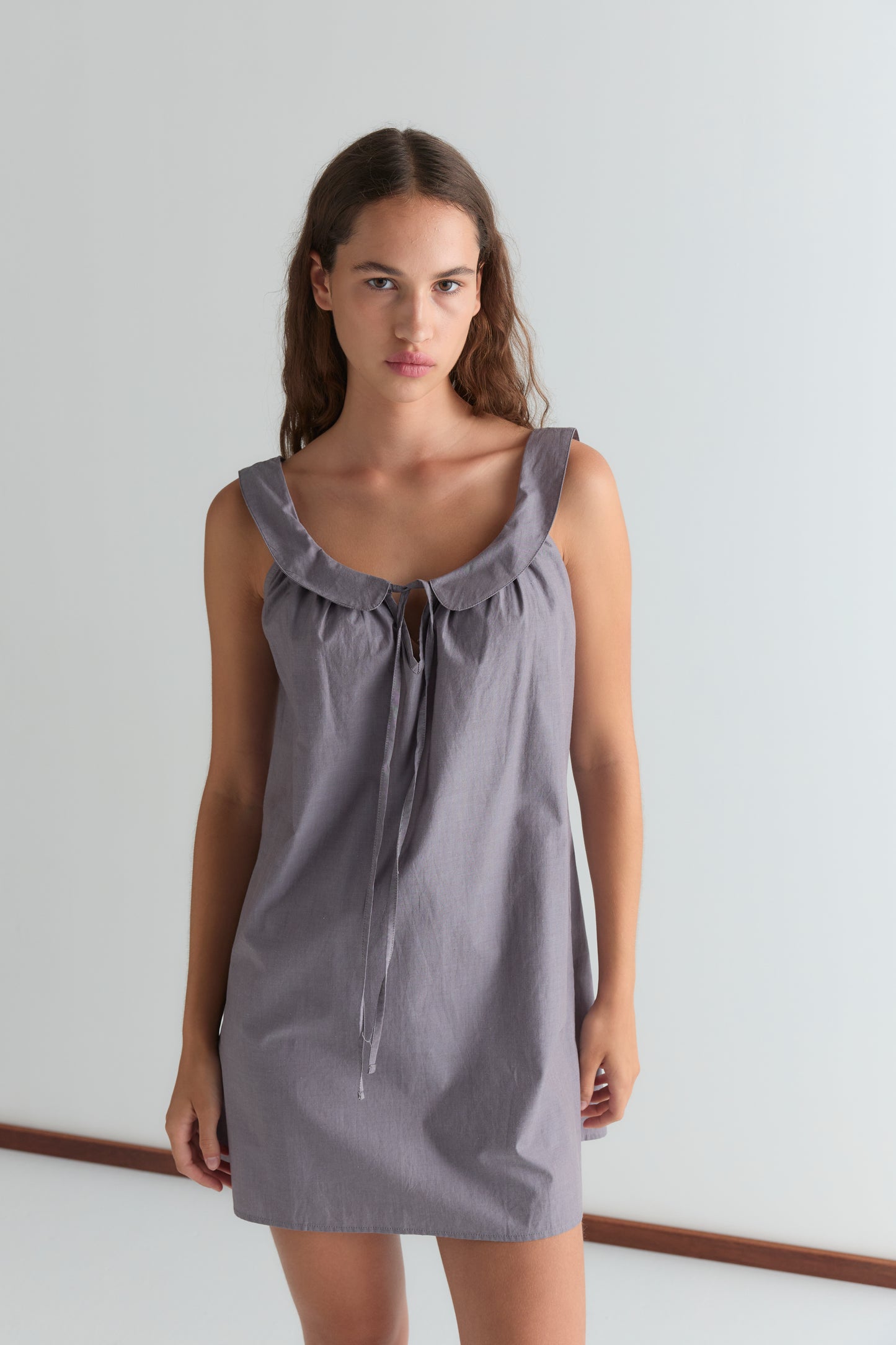 A woman stands against a white background, wearing the Rounded Neck Dress - Currant by Deiji Studios. The sleeveless dress features a ruffled neckline and front tie. She looks directly at the camera with a neutral expression.