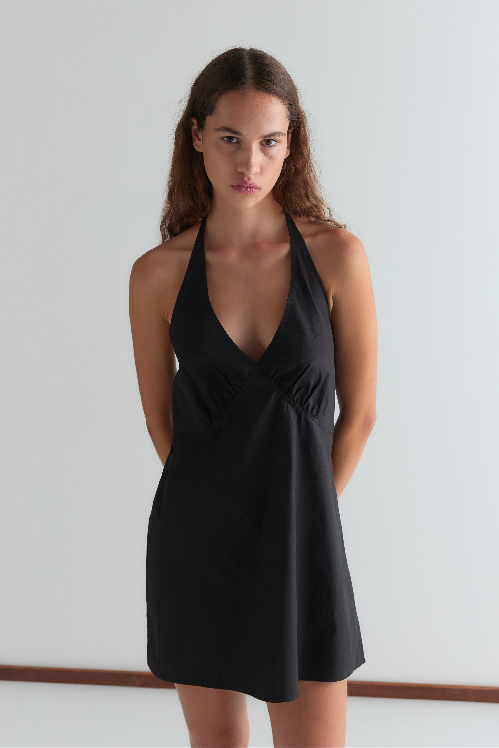 A woman with long brown hair stands against a plain light background, wearing the Cinched Halter Dress - Black. She looks directly at the camera with a neutral expression and keeps her arms behind her back.