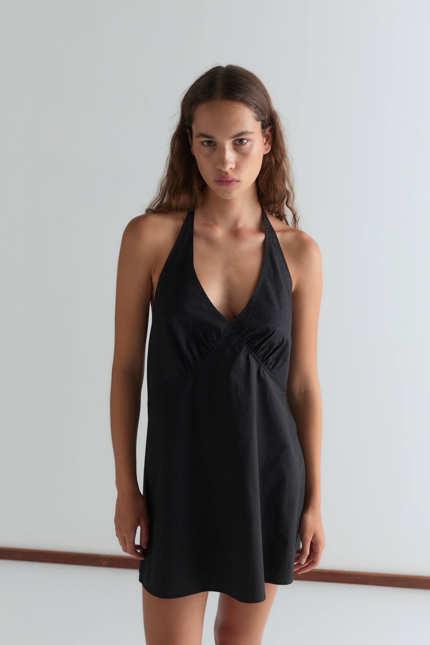 A woman with long, wavy brown hair models the Cinched Halter Dress - Black, sleeveless and halter-style, standing against a plain white background and looking directly at the camera with a neutral expression.