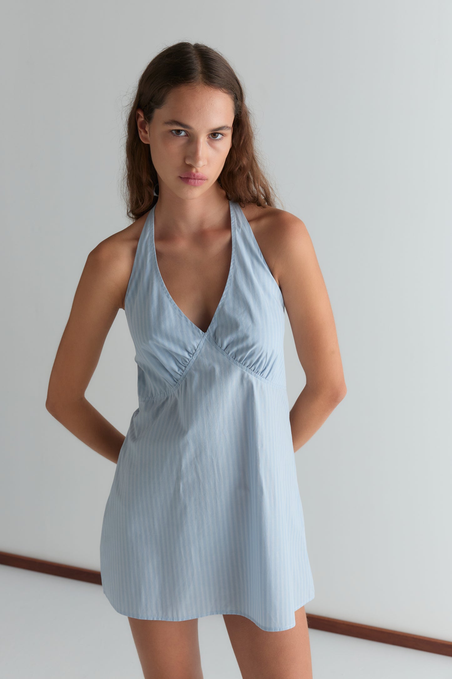 A woman with long brown hair stands indoors against a white wall, wearing the Cinched Halter Dress - Lake Blue Stripe. The dress features a halter neckline and subtle vertical stripes. She looks at the camera with her arms behind her back.