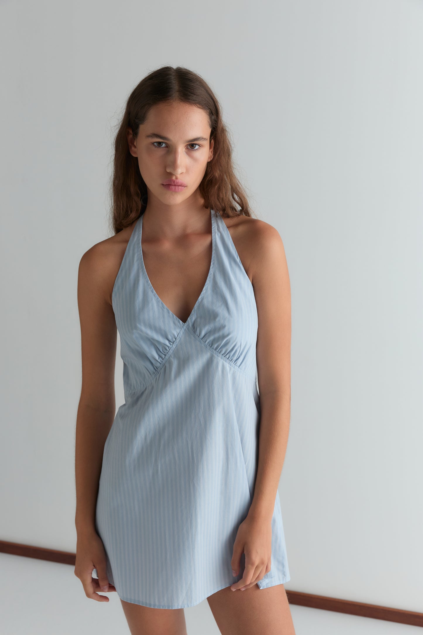 A woman with long brown hair wears the Cinched Halter Dress - Lake Blue Stripe, featuring a deep V-neckline, standing against a plain light background and looking at the camera with a neutral expression.