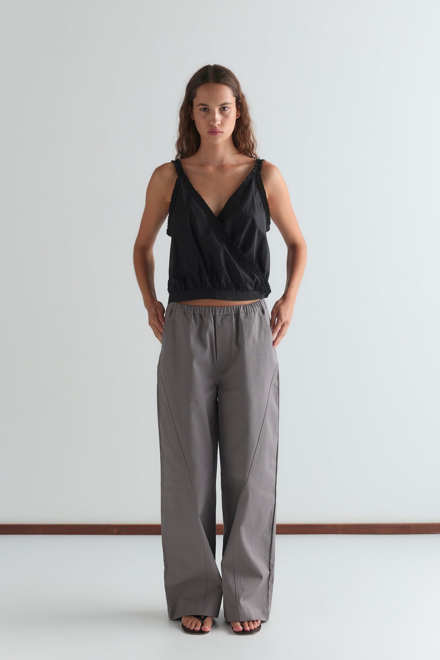 A woman stands against a plain white wall, wearing a sleeveless black wrap top and the Curve Pant in Lavender Smoke. The relaxed fit, mid-rise pants complement her look as she stands with hands in pockets, gazing directly at the camera.