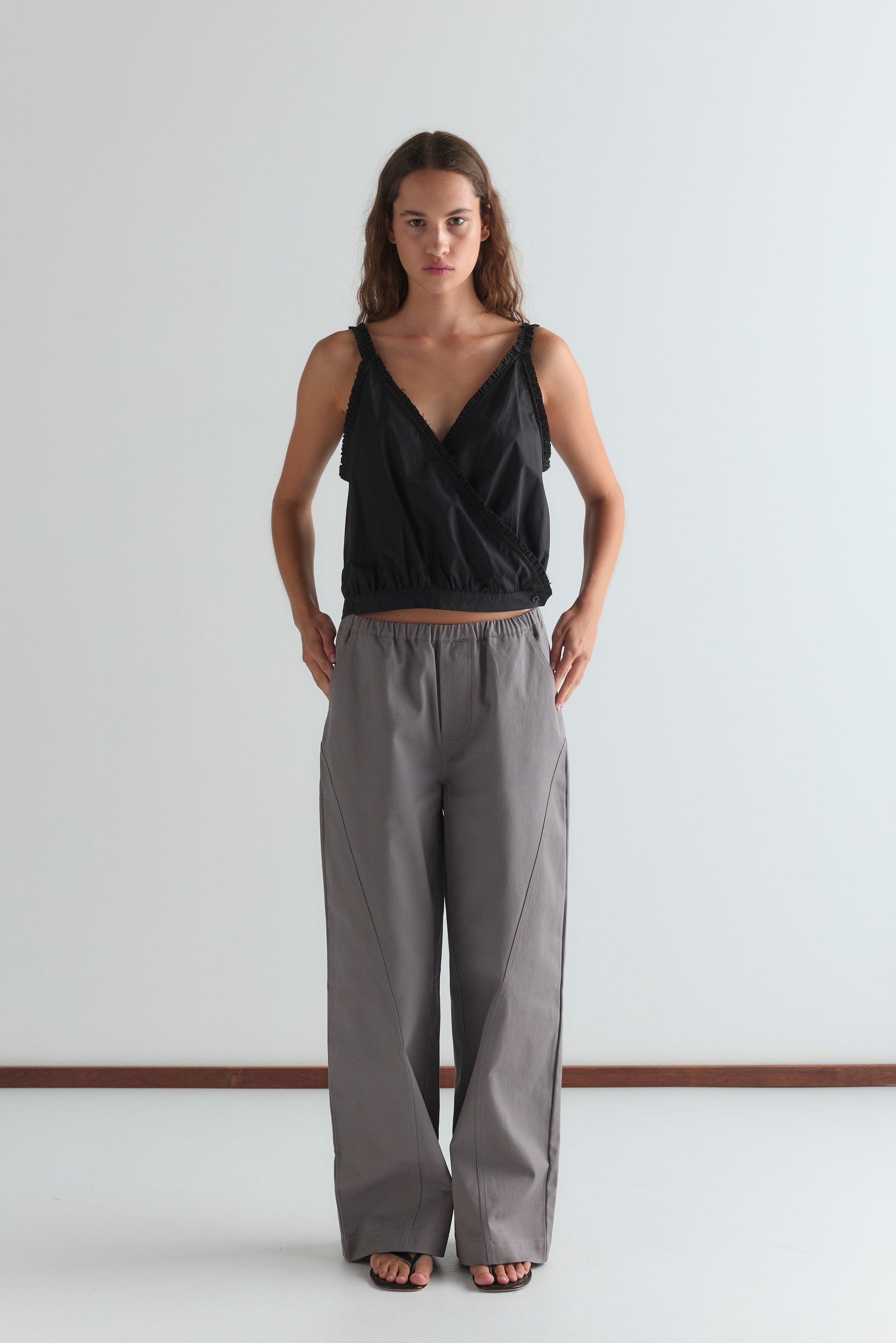 A woman stands against a plain white wall, wearing a sleeveless black wrap top and the Curve Pant in Lavender Smoke. The relaxed fit, mid-rise pants complement her look as she stands with hands in pockets, gazing directly at the camera.