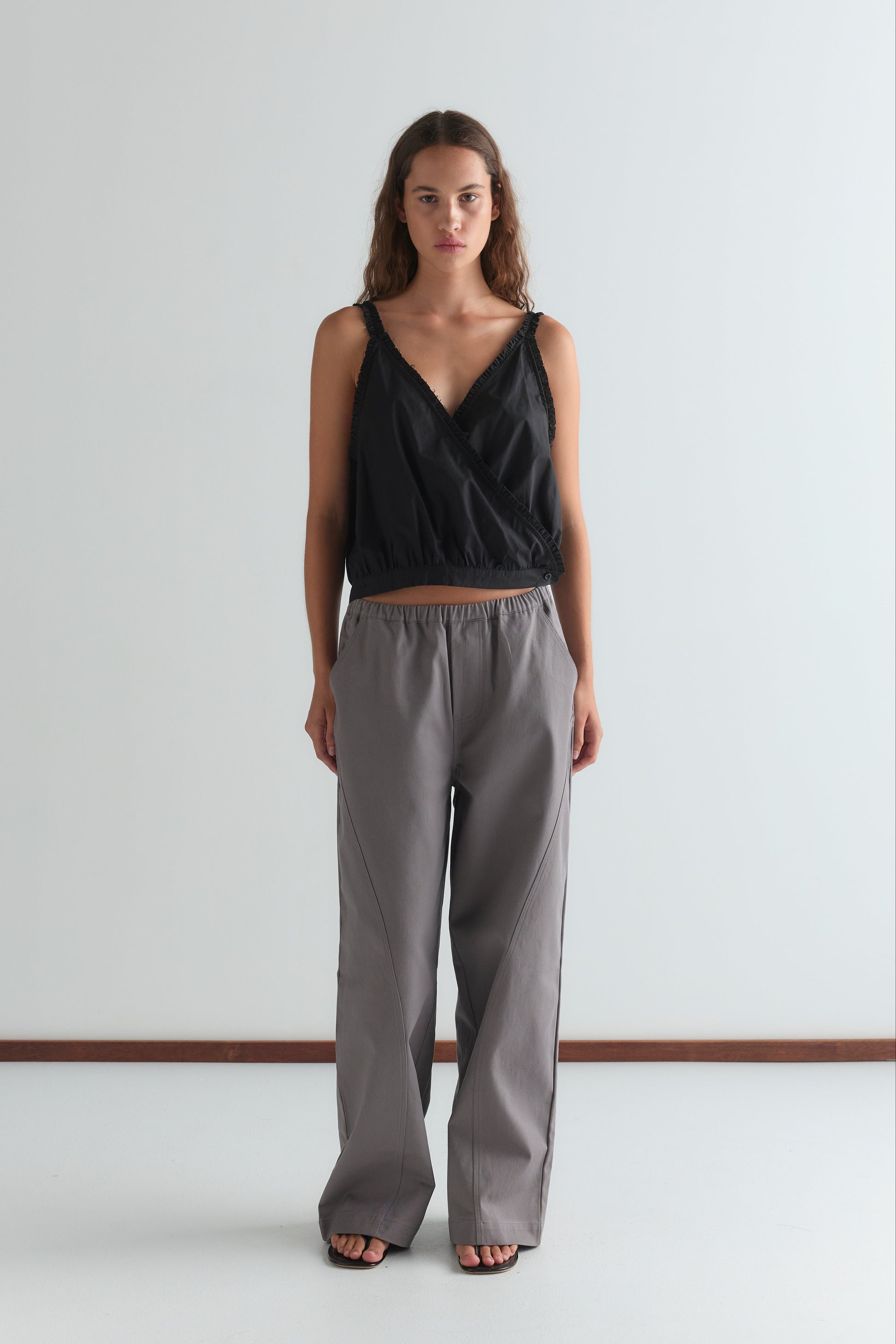 A woman faces forward in a sleeveless black wrap top and the Curve Pant - Lavender Smoke, featuring a relaxed fit and mid-rise waist, paired with black sandals against a plain white background.