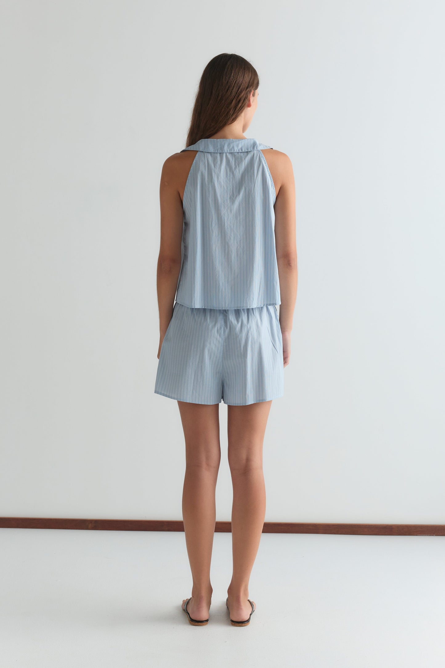 A woman stands barefoot on a white floor, wearing the Round Collar Set - Lake Blue Stripe: a light blue, sleeveless striped top and matching shorts by Deiji Studios. Her long hair drapes softly against a plain white wall.