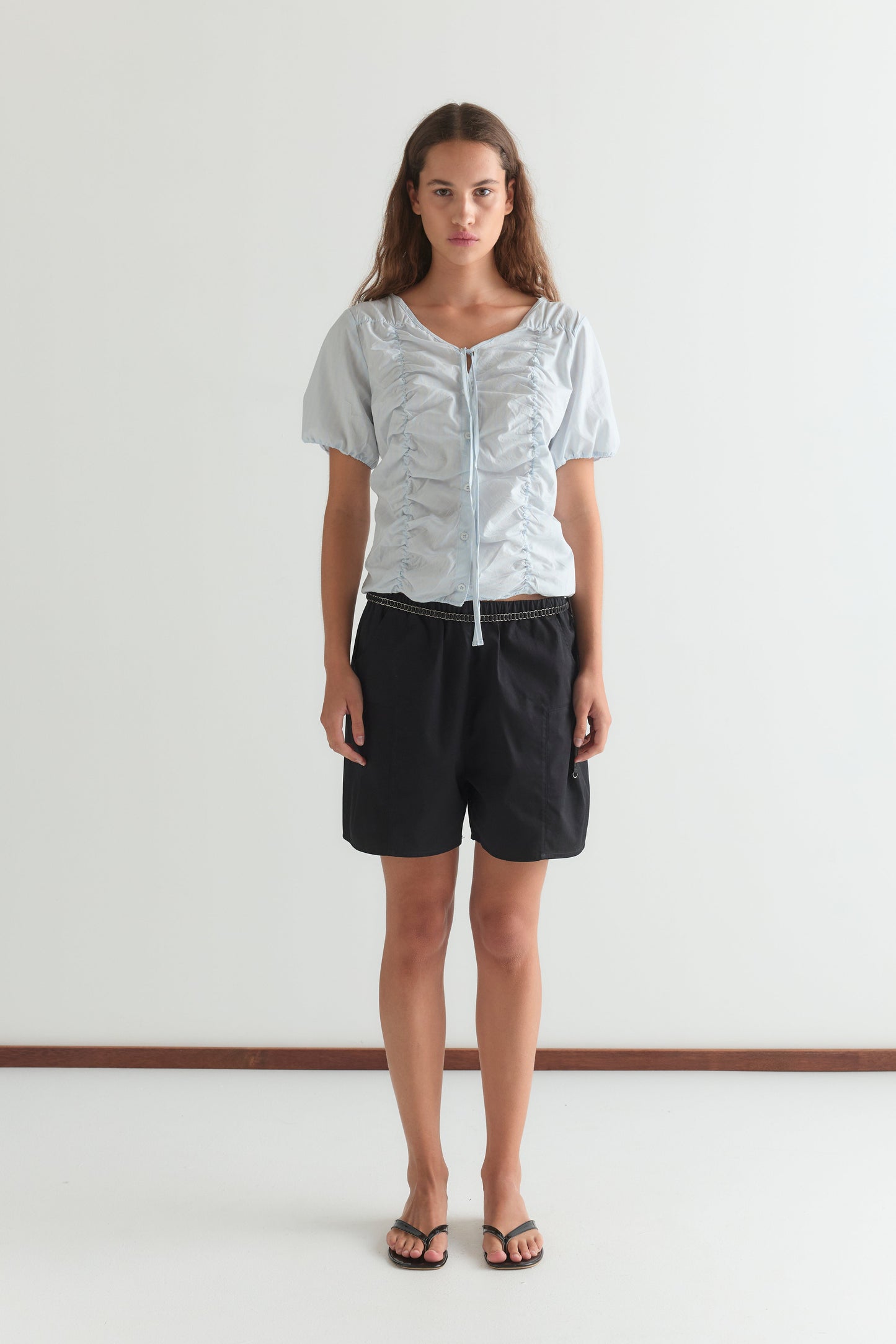 A woman with long brown hair and a neutral expression stands facing forward against a plain white wall, wearing the Crinkle Top - Powder Blue Stripe, black shorts, and black flip-flops.