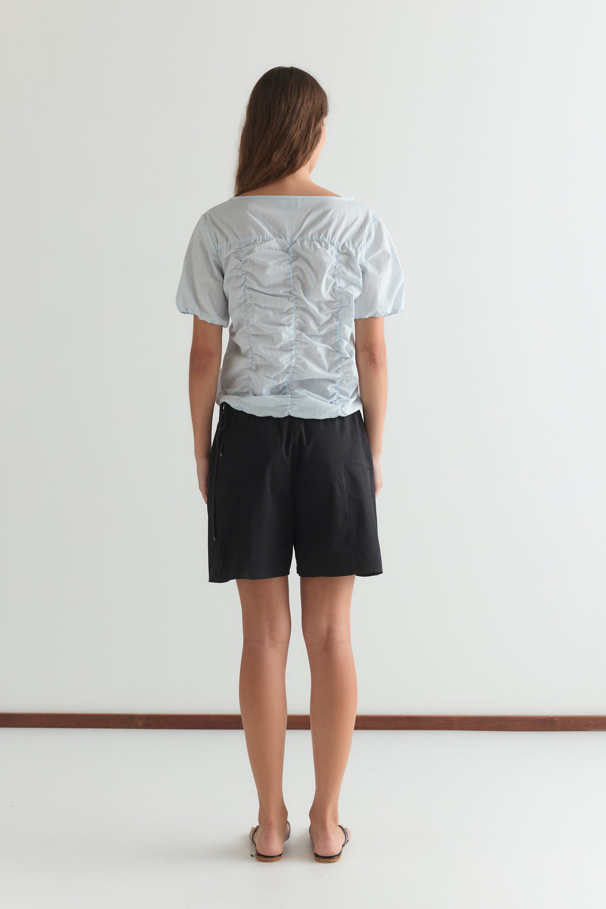 A woman with long brown hair, facing away, wears the Crinkle Top in Powder Blue Stripe with black shorts and sandals against a plain white background.