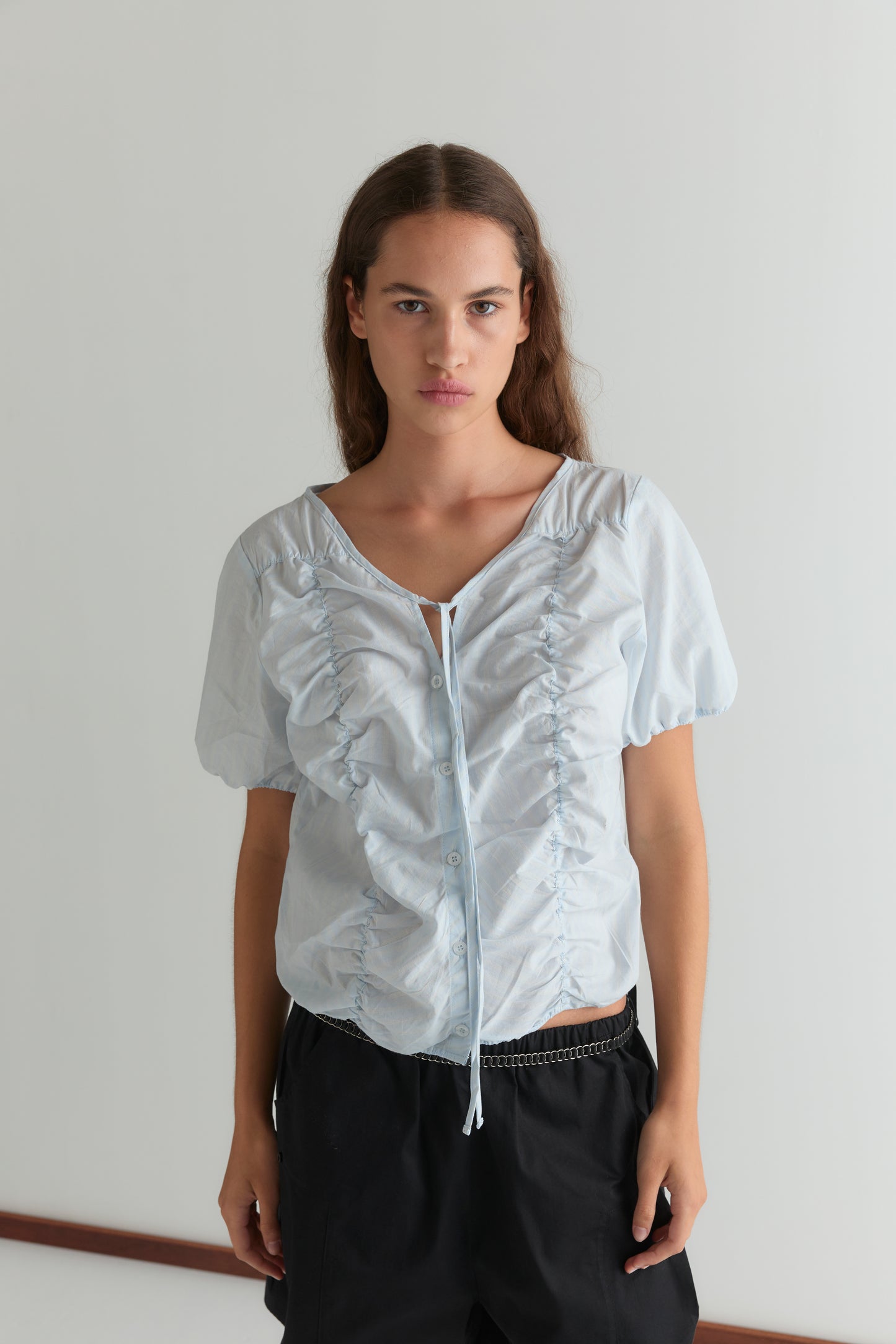 A woman with long brown hair wears the Crinkle Top - Powder Blue Stripe and black pants, standing against a plain white wall, looking directly at the camera with a neutral expression.
