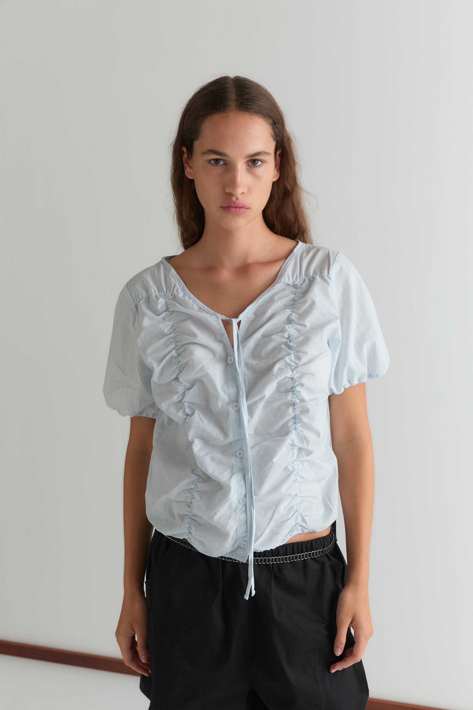 A woman with long brown hair wears the Crinkle Top - Powder Blue Stripe and black pants, standing against a plain white wall, looking directly at the camera with a neutral expression.