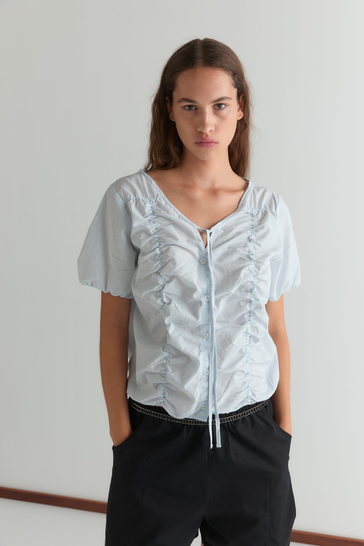A woman with long brown hair stands against a white background, wearing the Crinkle Top in powder blue stripe with short puff sleeves and black pants, her hands in her pockets.