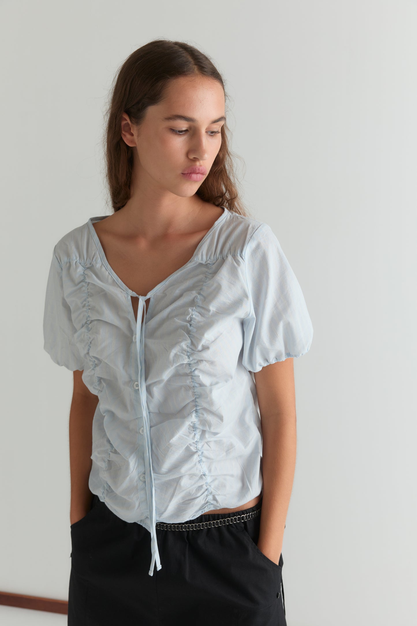 A woman with long brown hair wears the Crinkle Top - Powder Blue Stripe, featuring lightweight sheer fabric, ruched details, and short puff sleeves. She pairs it with black pants and stands against a white background, looking down with a neutral expression.
