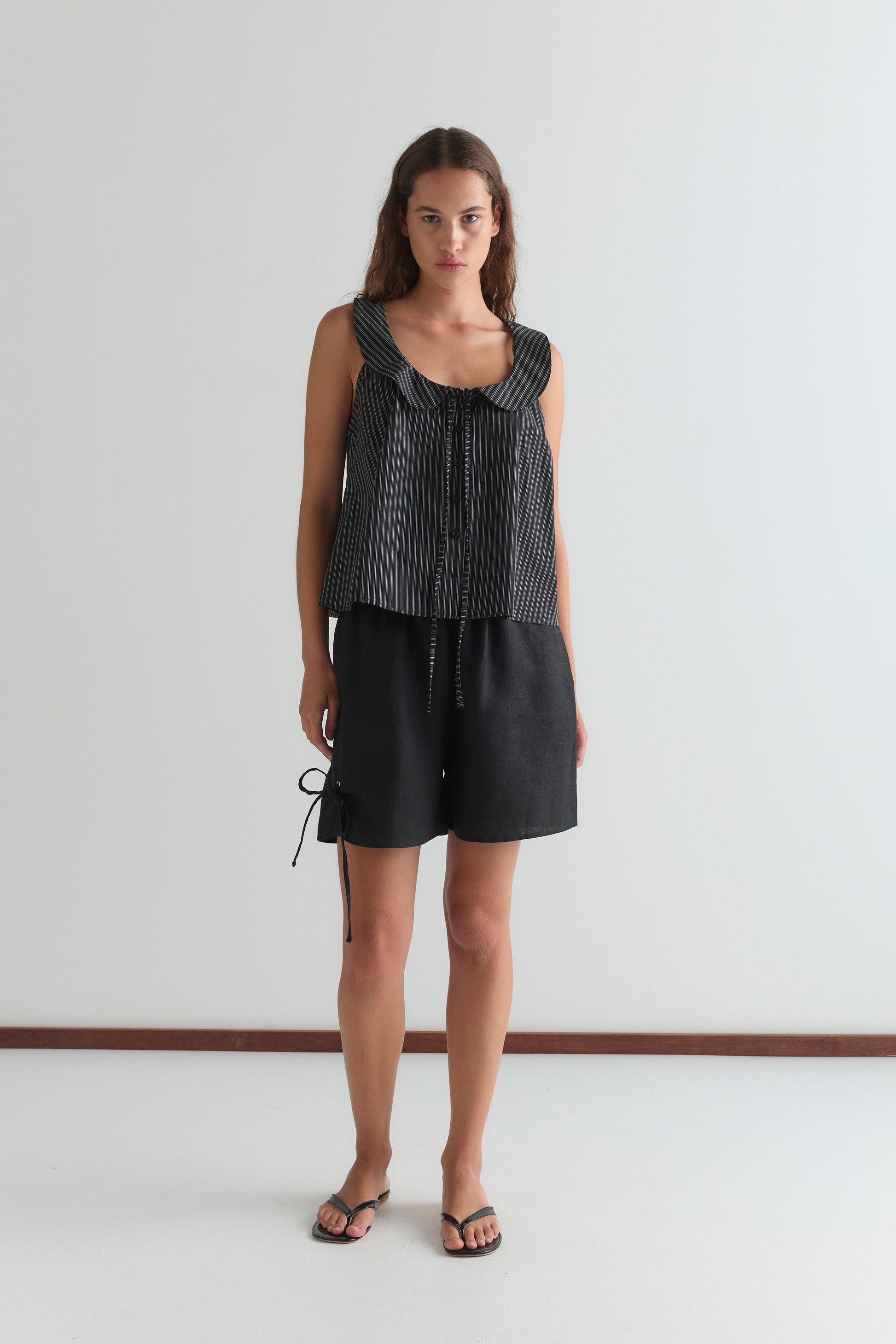A woman with long brown hair and a neutral expression stands against a white wall, wearing a Deiji Studios sleeveless black and white striped tie-front top, Eyelet Short - Black with side tie detail, and black sandals.