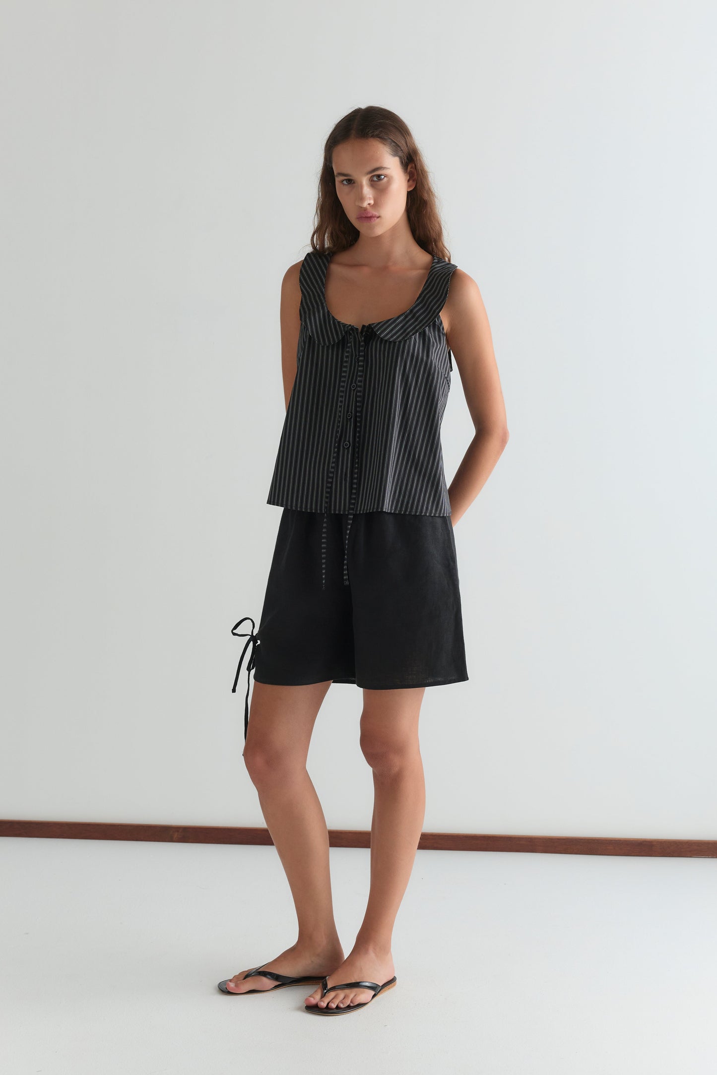 A woman stands against a white wall wearing the Round Collar Top - Mono Stripe with a front tie, black side-tie shorts, and black sandals. Her long hair is loose, and she has a neutral expression.