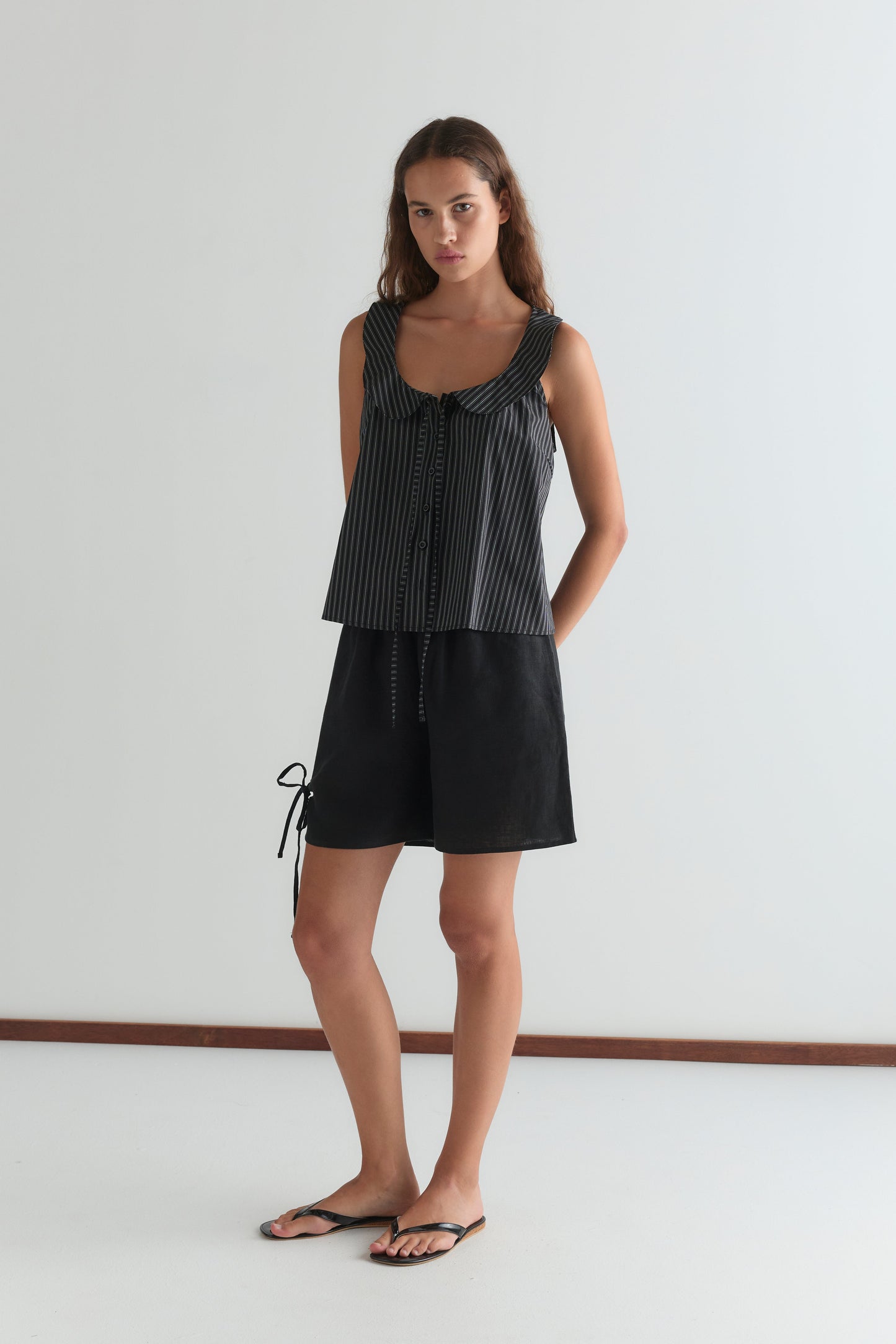 A woman stands against a plain white wall, wearing a sleeveless black and white patterned top, Deiji Studios Eyelet Short - Black with side ties, and black flip-flops. She has long brown hair and a neutral expression, hands behind her back.