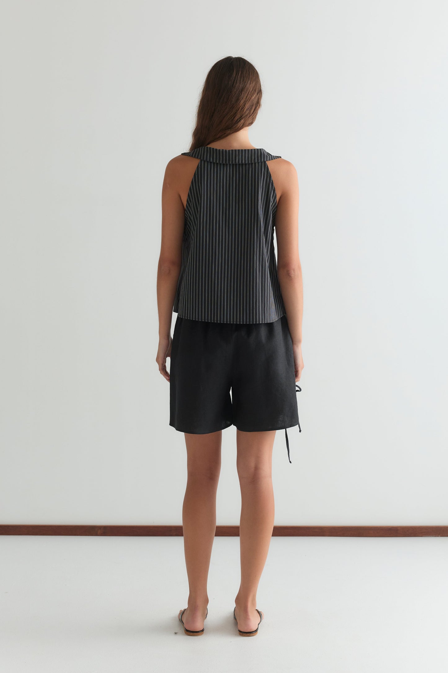 A woman with long brown hair stands barefoot, facing away, in a Deiji Studios Round Collar Top - Mono Stripe with black and gray stripes and loose black shorts, against a plain white wall and floor background.