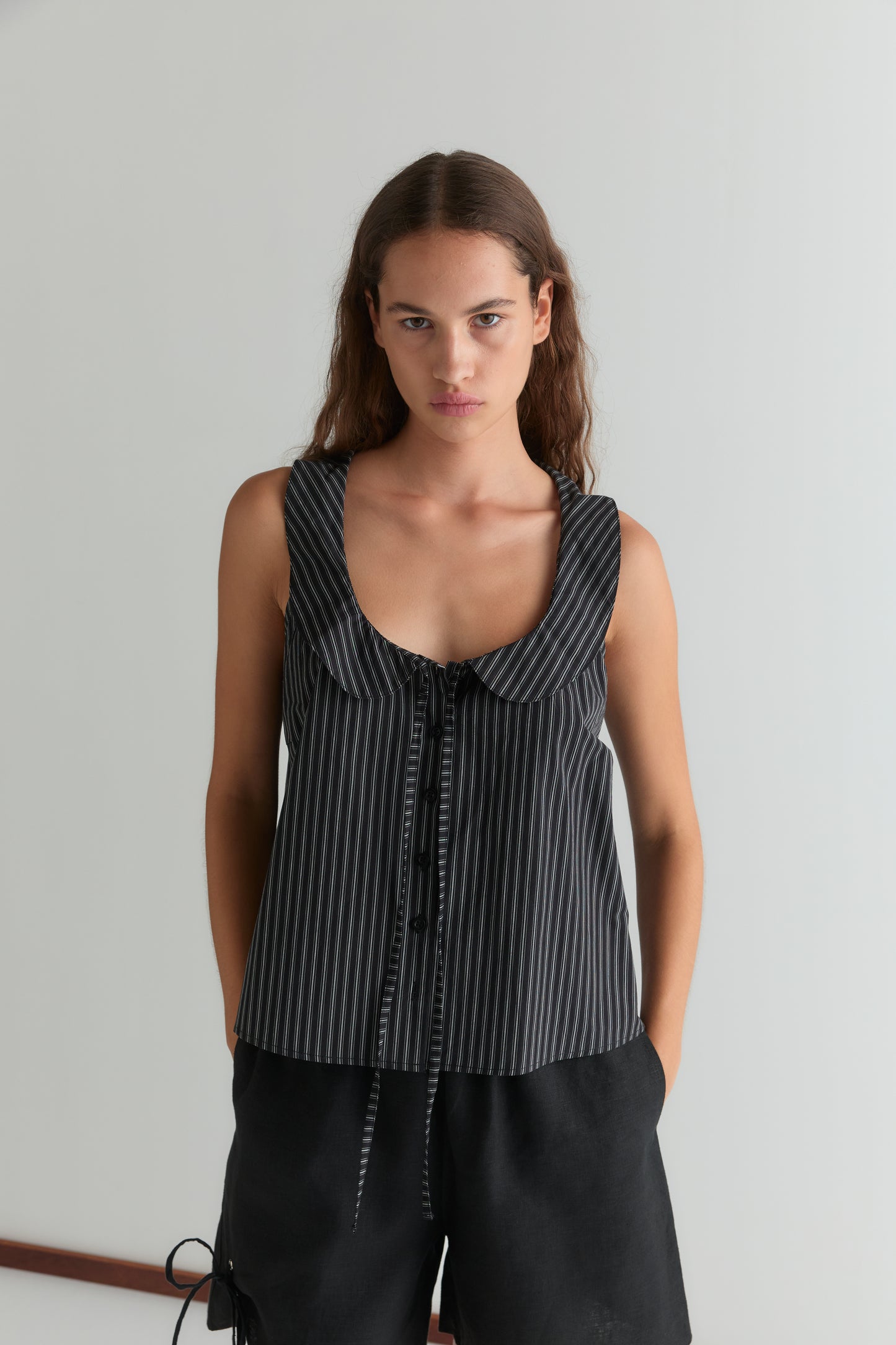 A woman with long brown hair wears the Deiji Studios Round Collar Top - Mono Stripe, a sleeveless piece with black and gray stripes, rounded collar, and bow tie detail, paired with black shorts against a plain light background.
