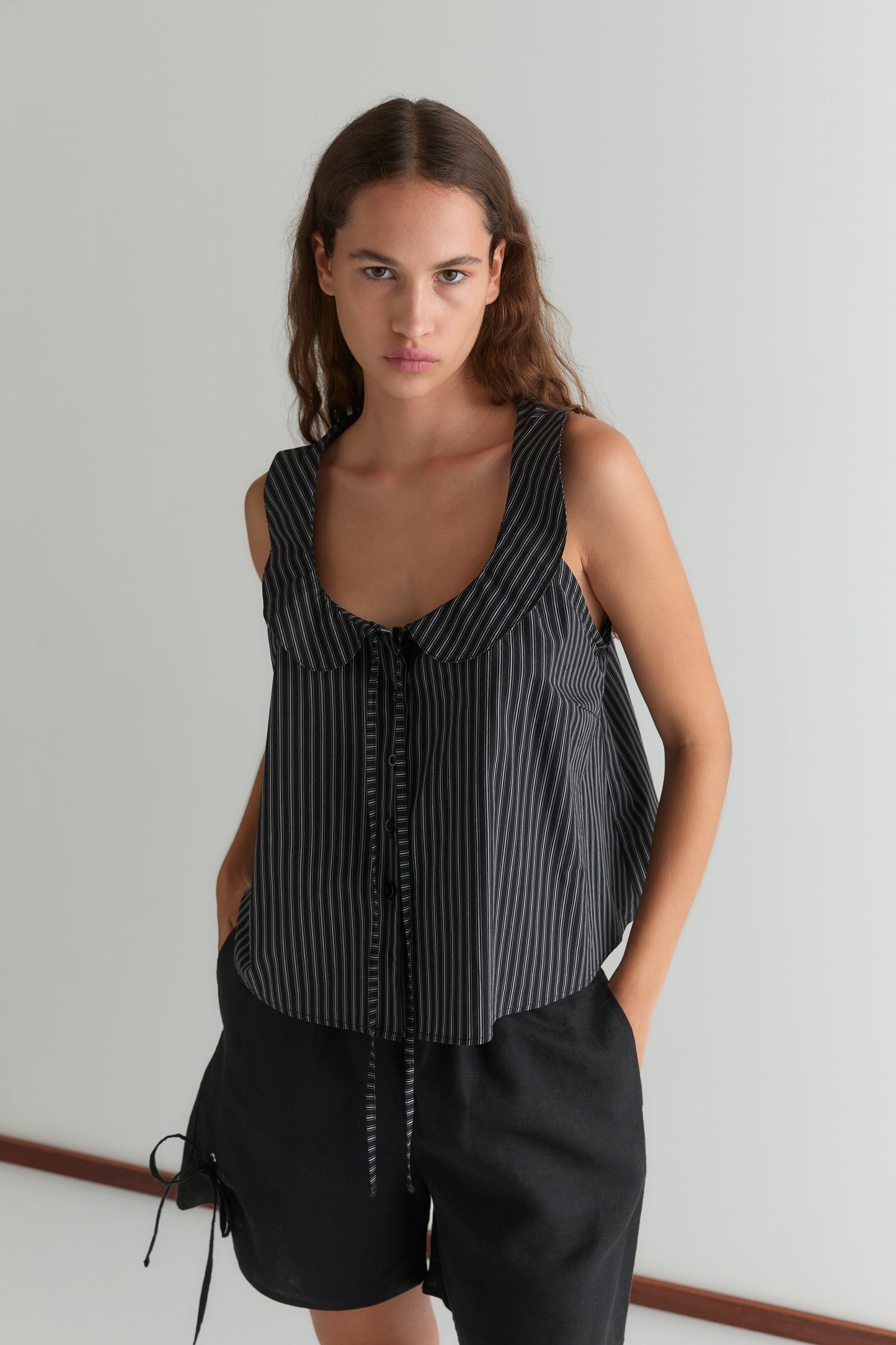 A woman with long brown hair wears a sleeveless, black and white pinstriped front-tie top and Eyelet Short - Black, standing against a plain white wall with her hands in her pockets.