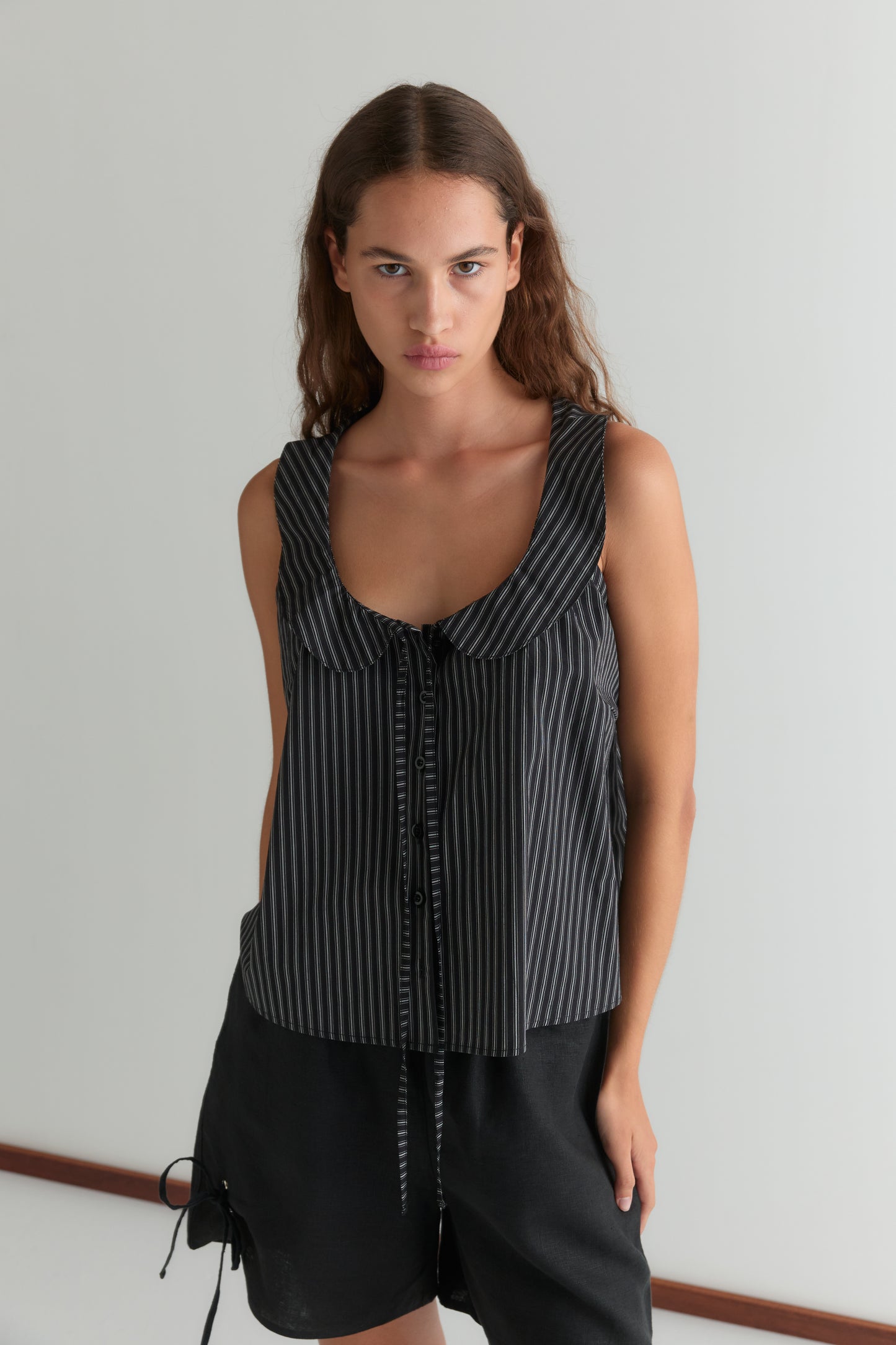 A woman with long, wavy brown hair stands against a plain light background, wearing the Round Collar Top - Mono Stripe by Deiji Studios and black shorts. She looks directly at the camera with a neutral expression.