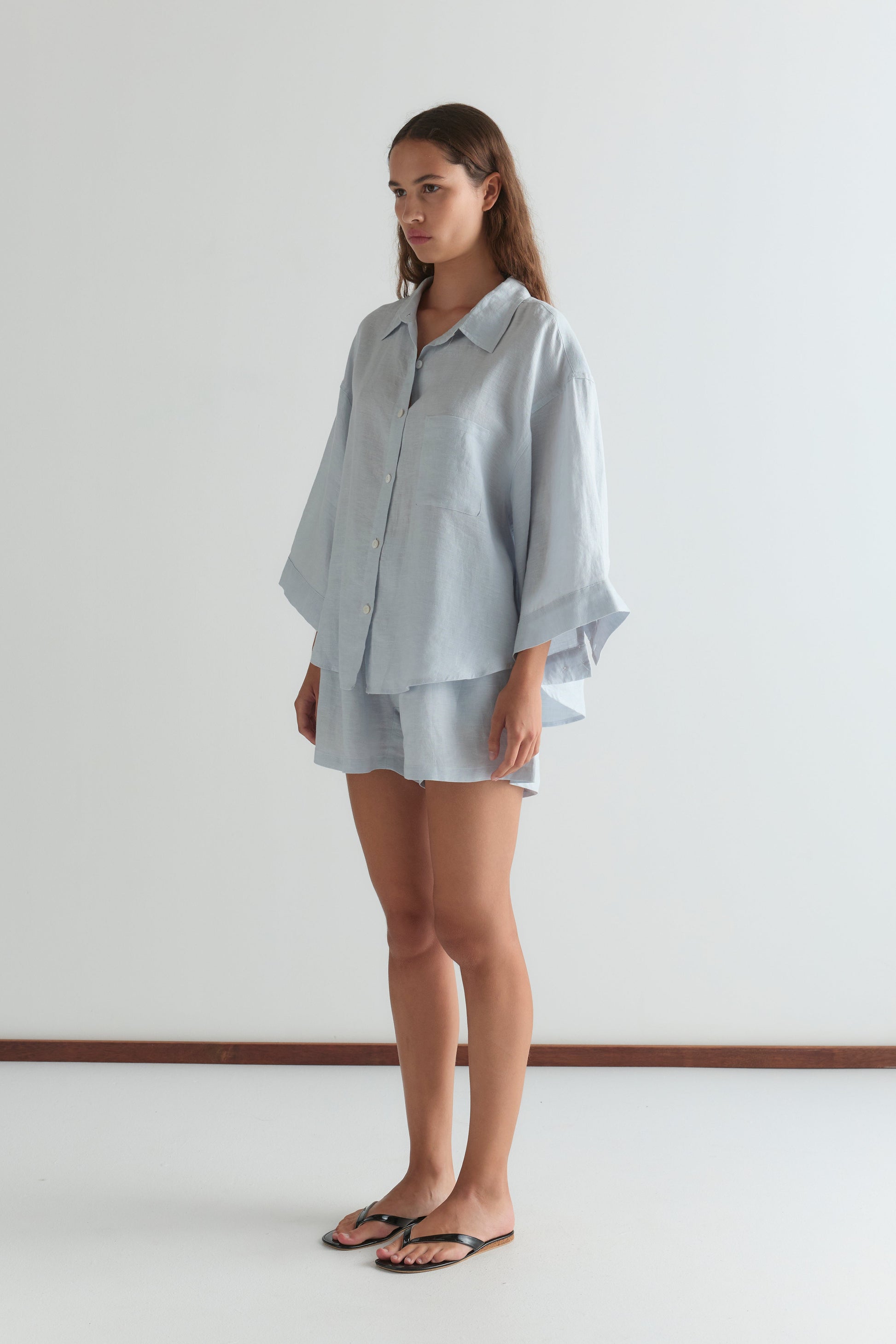 A woman with long brown hair stands indoors against a plain white wall, wearing Deiji Studios' 03 Set in Soft Blue—an oversized linen shirt with wide sleeves and matching boxer-style shorts—paired with black flip-flops.