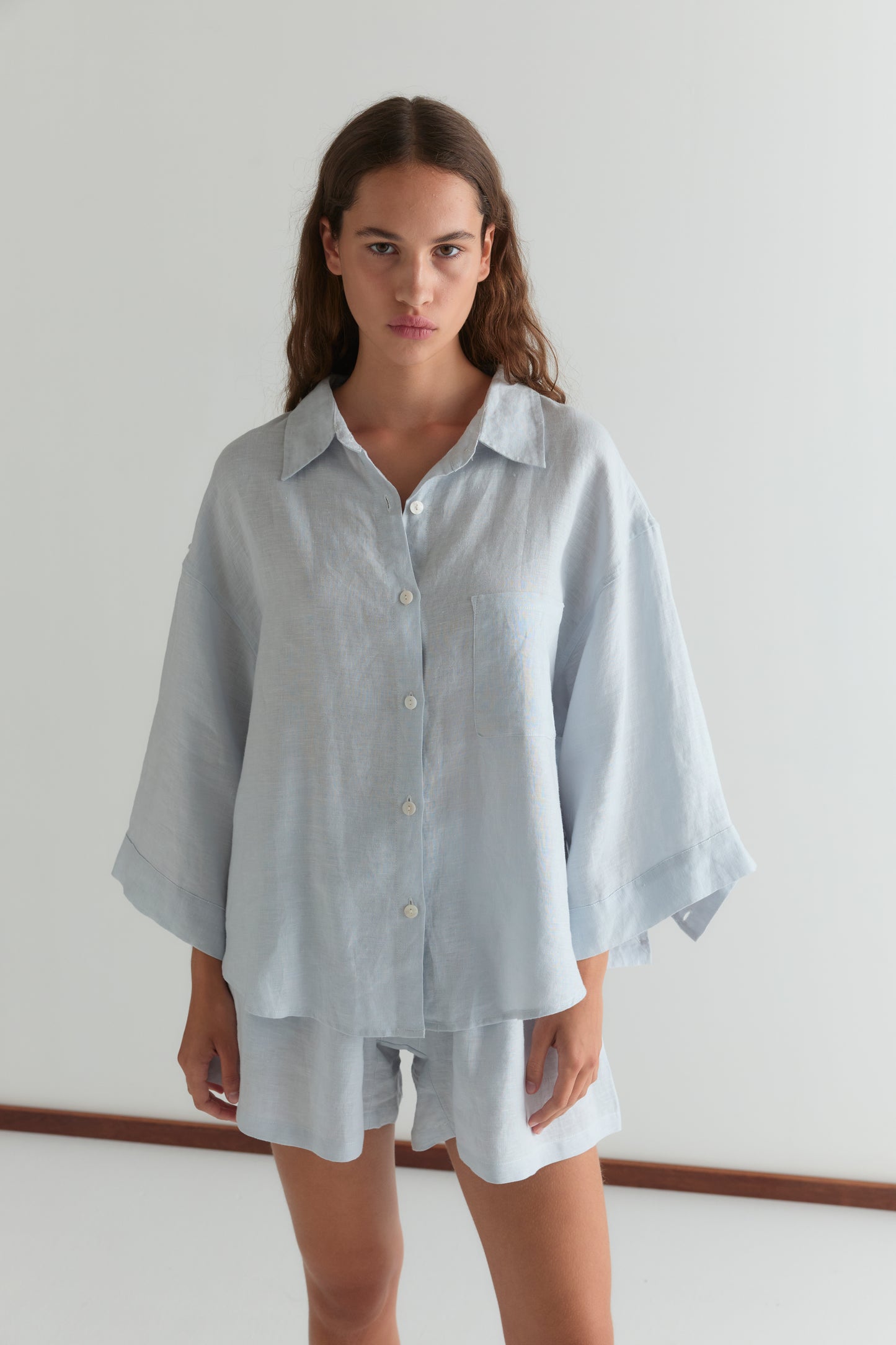 A woman with long brown hair stands against a white background, wearing the 03 Set - Soft Blue: an oversized linen shirt and matching boxer style shorts. She looks directly at the camera with a neutral expression.