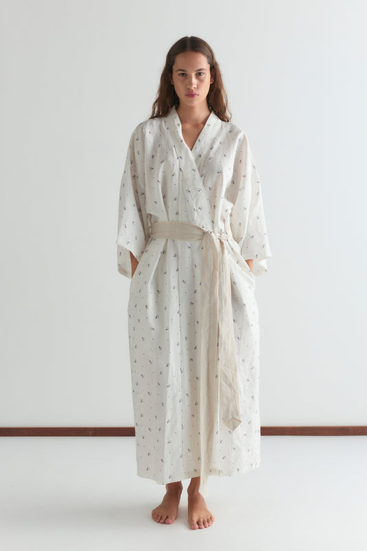 A woman stands barefoot against a plain background, wearing the 02 Robe—a long, loose white robe with a subtle pattern and beige sash at the waist. She faces forward with her hands in the robe’s pockets.