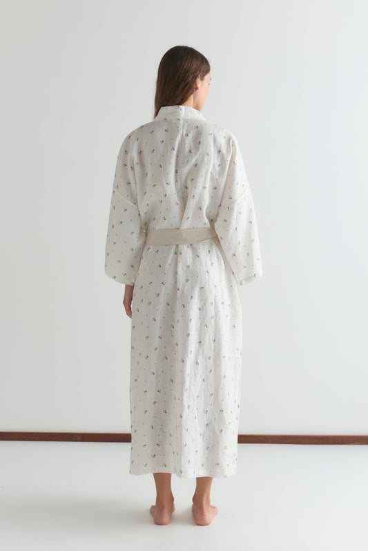 A person with long brown hair stands barefoot and facing away, wearing the 02 robe - vintage floral, which is ankle-length, light-colored, features a linen waist tie, and styled against a plain white background.