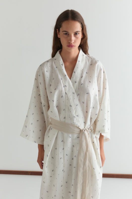 A woman with long, wavy brown hair and a neutral expression stands against a plain white background, wearing the 02 robe - vintage floral, a full-length style featuring a linen waist tie and a subtle dotted pattern.