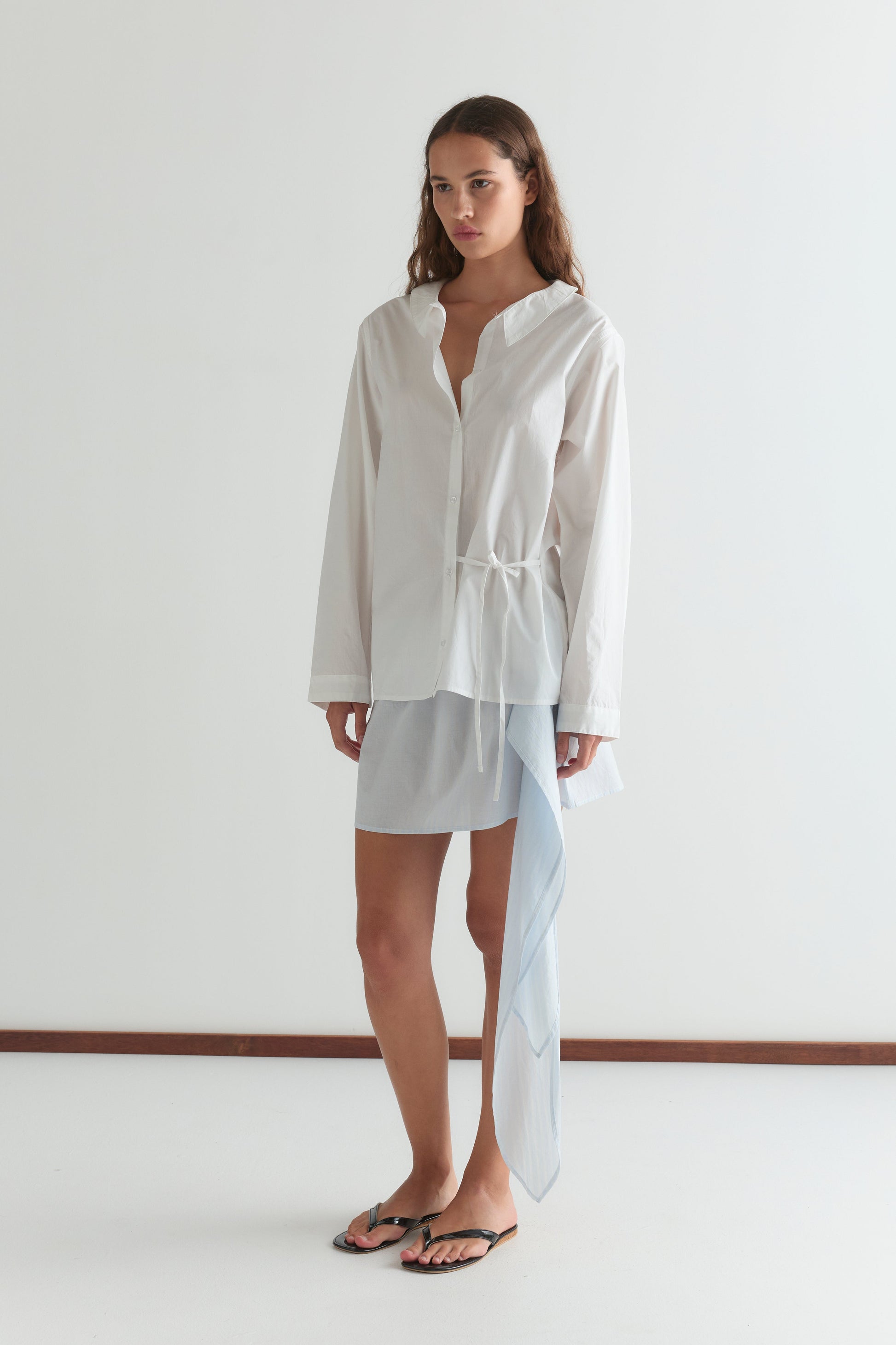 A woman with long, wavy brown hair and a neutral expression stands indoors against a white wall, wearing the Left Shirt - White in a relaxed fit over a light blue skirt with an asymmetrical sheer panel and black sandals.