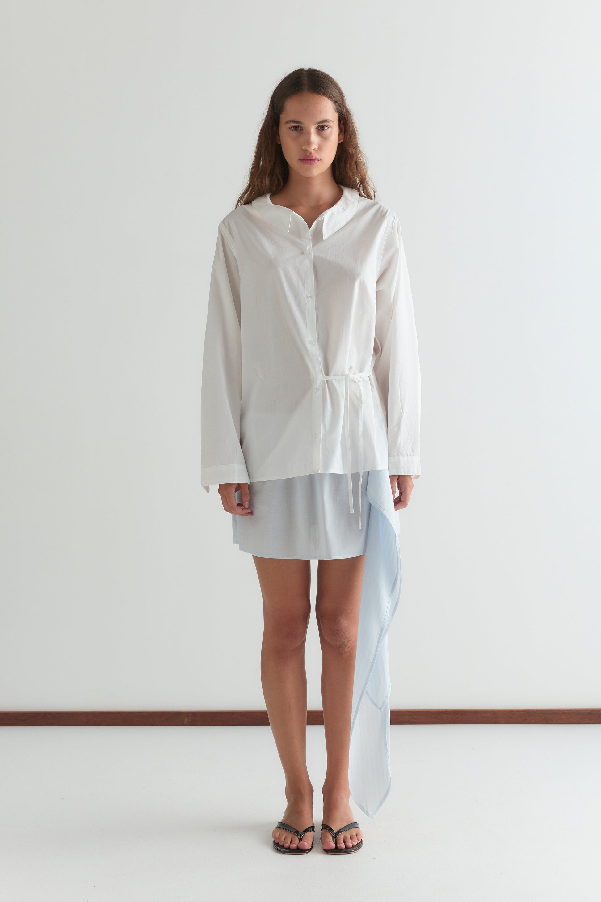 A woman with long, wavy hair and a neutral expression wears the Left Shirt in white against a plain background, paired with a short light blue skirt featuring an asymmetrical draped panel, adjustable side tie, and black sandals.