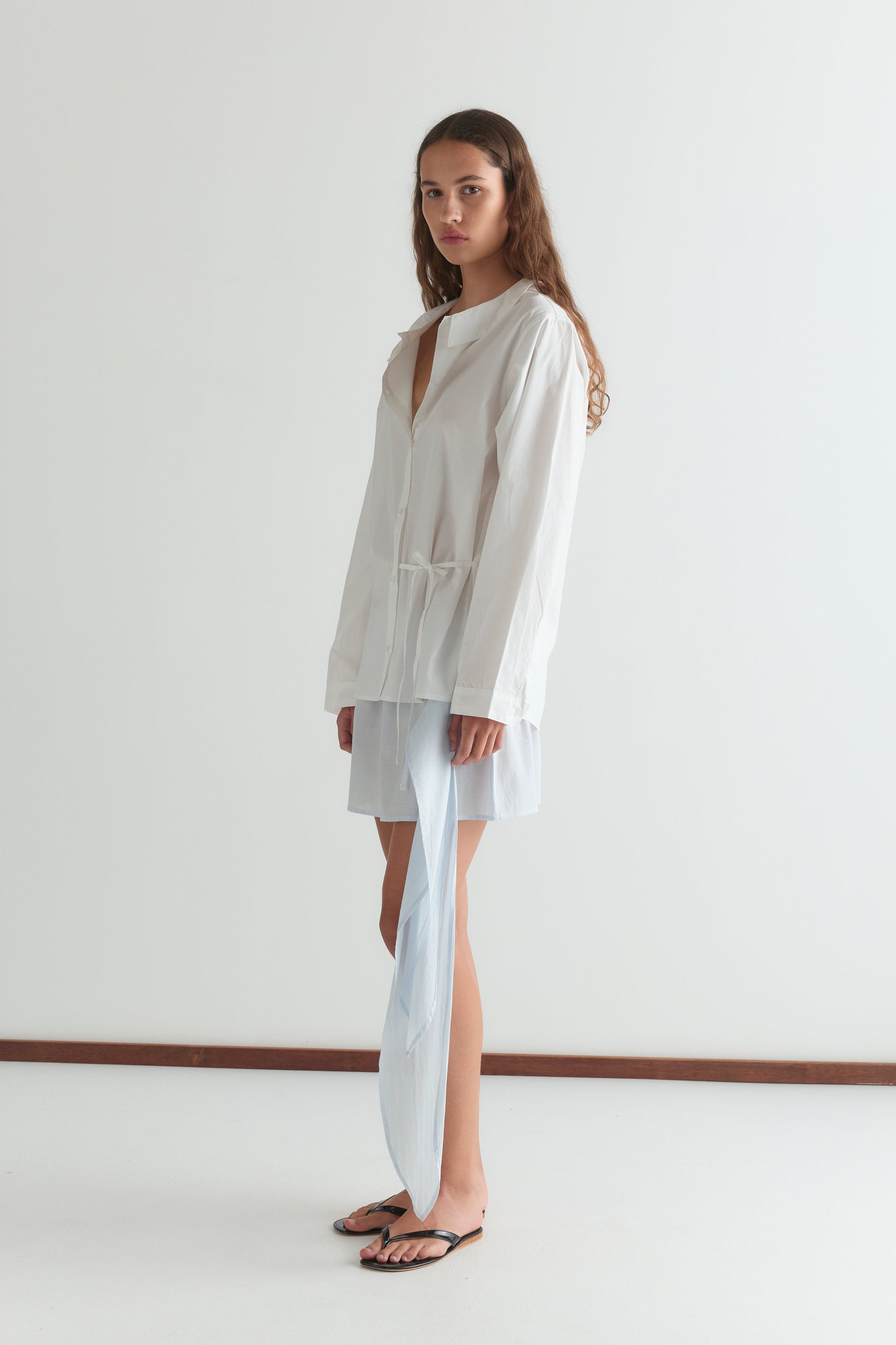 A woman with long brown hair stands against a white wall wearing the Left Shirt - White in a relaxed silhouette, paired with a light blue skirt or shorts and black flip-flops. She gazes forward with a neutral expression.