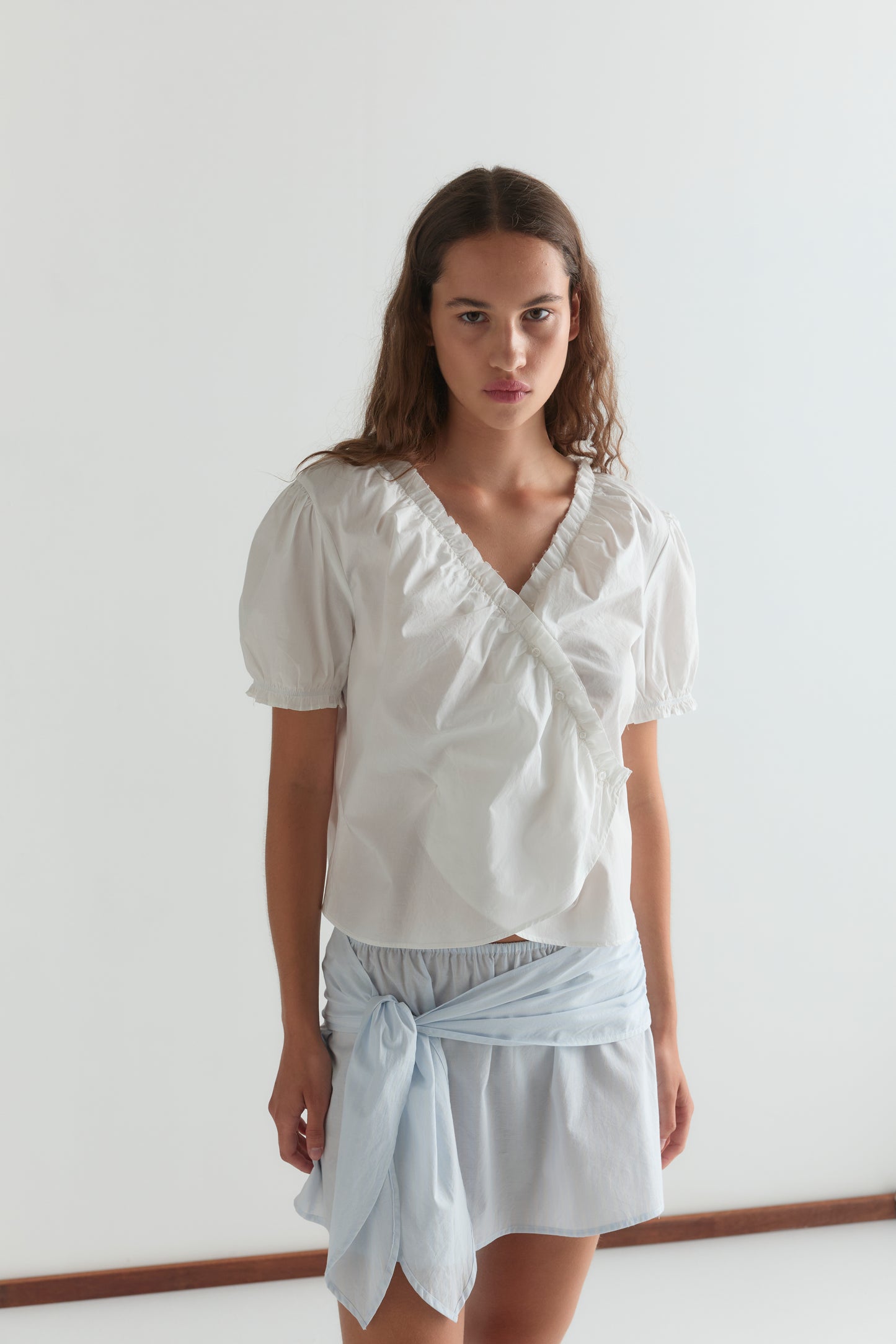 A woman with long brown hair wears a white puff-sleeve wrap blouse and a Belted Skirt - Powder Blue Stripe from Deiji Studios, styled with a tied sash, standing against a plain white wall.