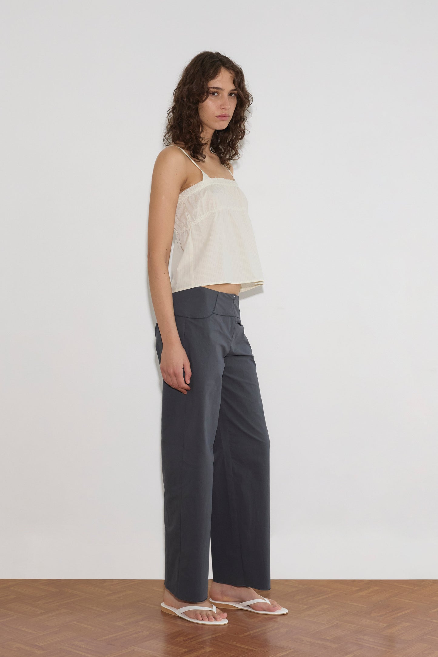 A woman with curly hair stands against a plain white wall, wearing the Tri Cami - Citrus Stripe from Deiji Studios, paired with loose dark gray pants and white sandals. She gazes slightly to the side with a neutral expression.