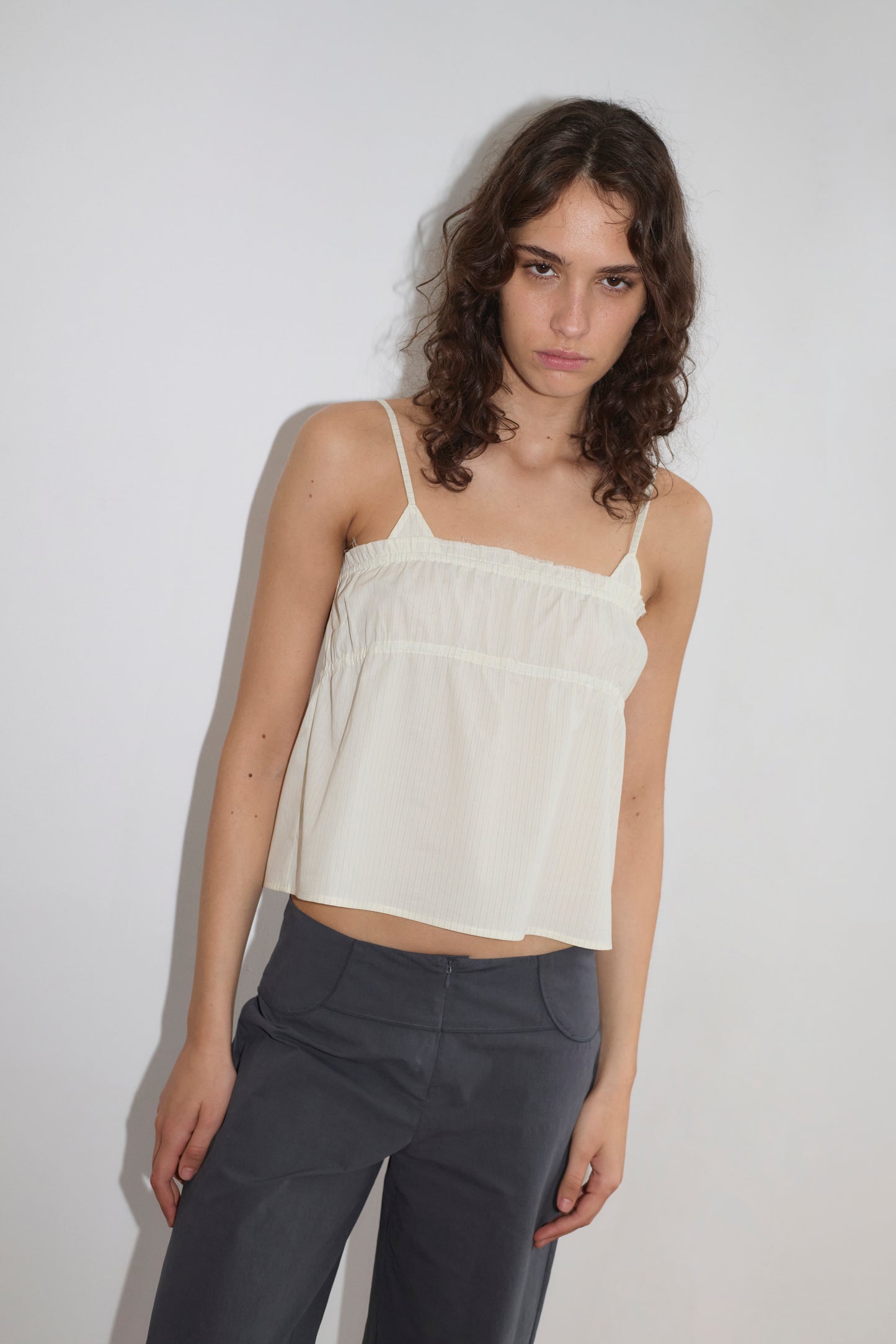 A young woman with wavy brown hair stands against a plain white background, wearing the Tri Cami in Citrus Stripe by Deiji Studios and dark gray pants. She looks slightly downward with a neutral expression.