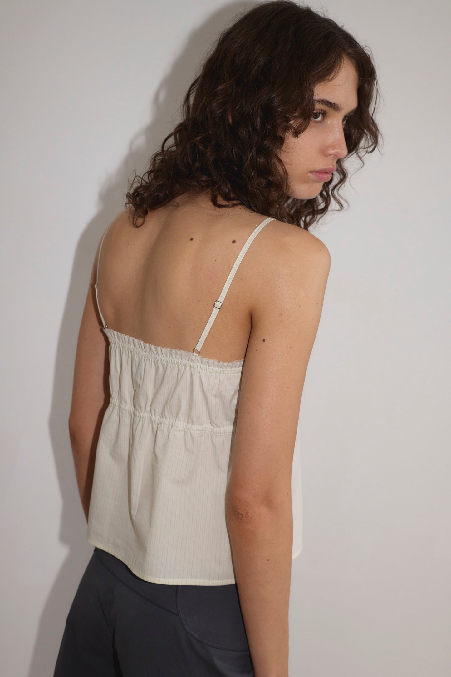 A person with curly brown hair wears the Tri Cami - Citrus Stripe by Deiji Studios with dark pants, standing against a plain white background and looking over their shoulder toward the camera.