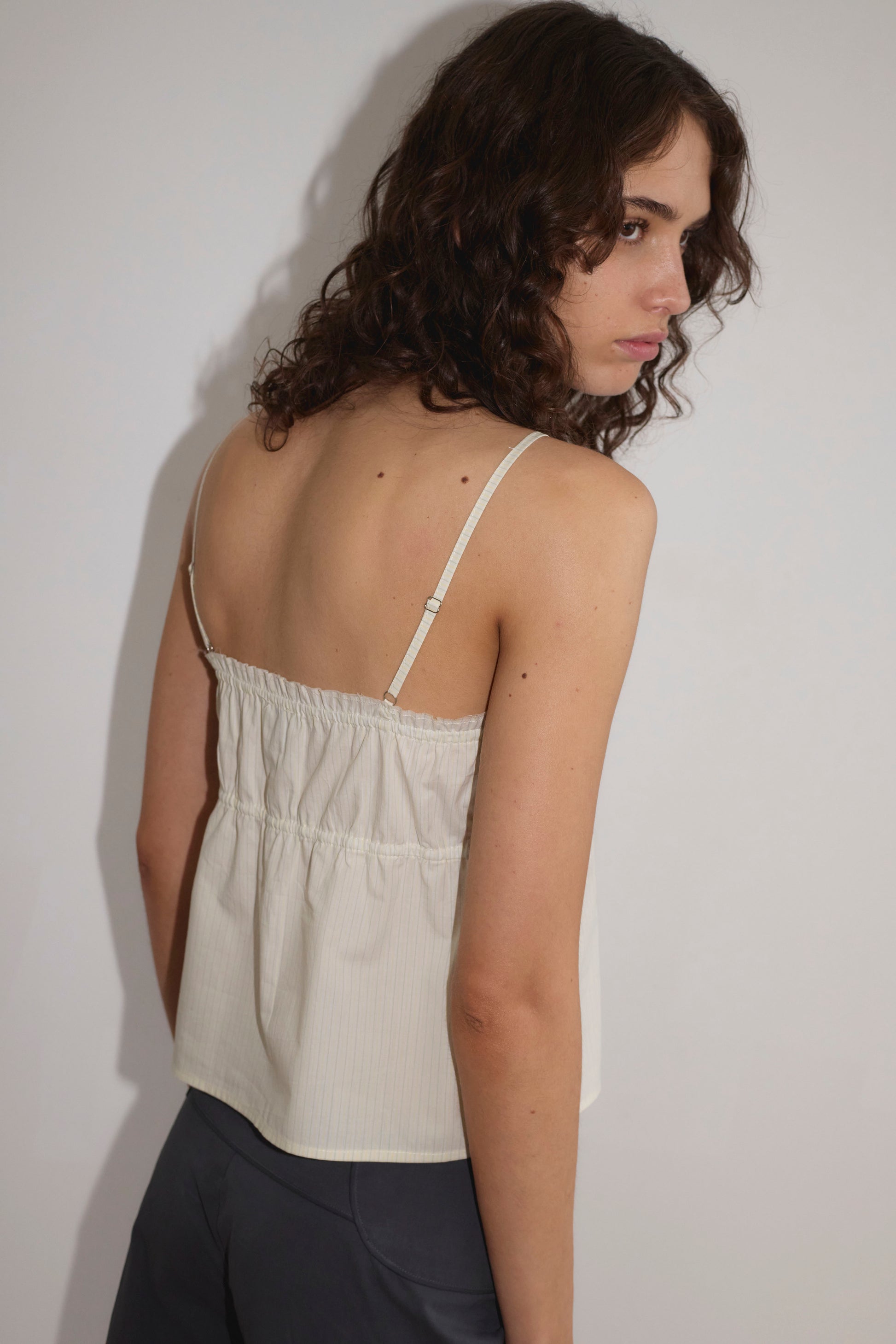 A person with curly brown hair wears the Tri Cami - Citrus Stripe by Deiji Studios with dark pants, standing against a plain white background and looking over their shoulder toward the camera.