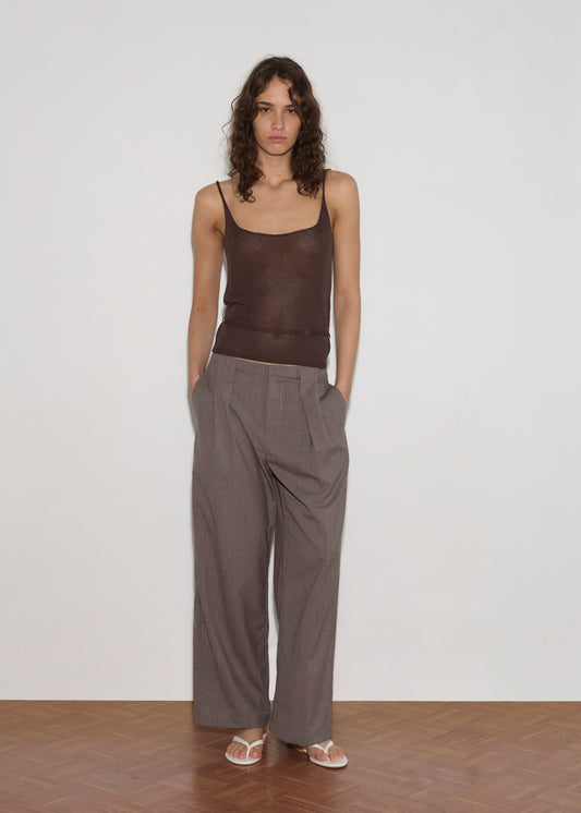 A person with long curly hair stands against a plain white wall, wearing a Square Knit Top - Brown, wide-legged gray plaid trousers, and white sandals on a wooden floor.