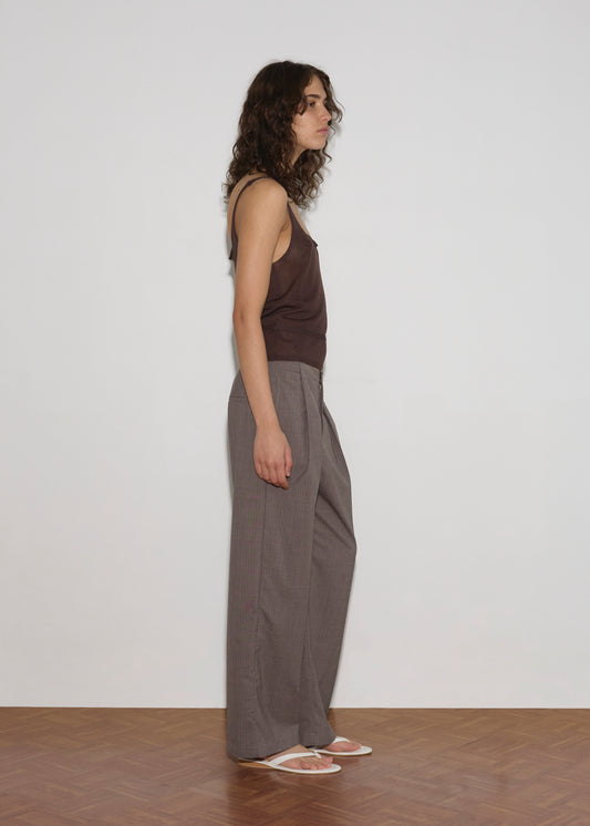 A person with curly brown hair stands in profile against a plain white wall, wearing a Square Knit Top - Brown, loose gray pants, and white flip-flops on a wooden floor.