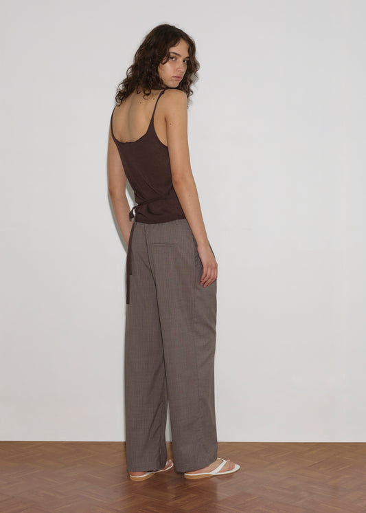 A woman with wavy brown hair wears the Square Knit Top - Brown by Deiji Studios, loose gray pants, and white sandals, standing on a wooden floor and looking over her shoulder against a plain white wall.