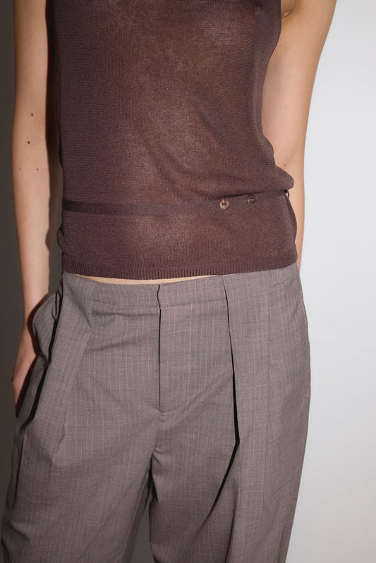 A person wears the Square Knit Top - Brown, a sheer fine knit tank, paired with high-waisted pleated grey trousers. Standing against a plain light background, one hand is in their pocket. The image is cropped from shoulders to knees.