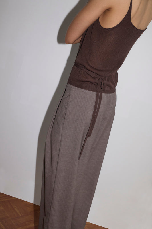 A person in a Deiji Studios Square Knit Top - Brown and loose, high-waisted gray trousers stands with arms crossed, facing away from the camera against a plain white wall and wooden floor.