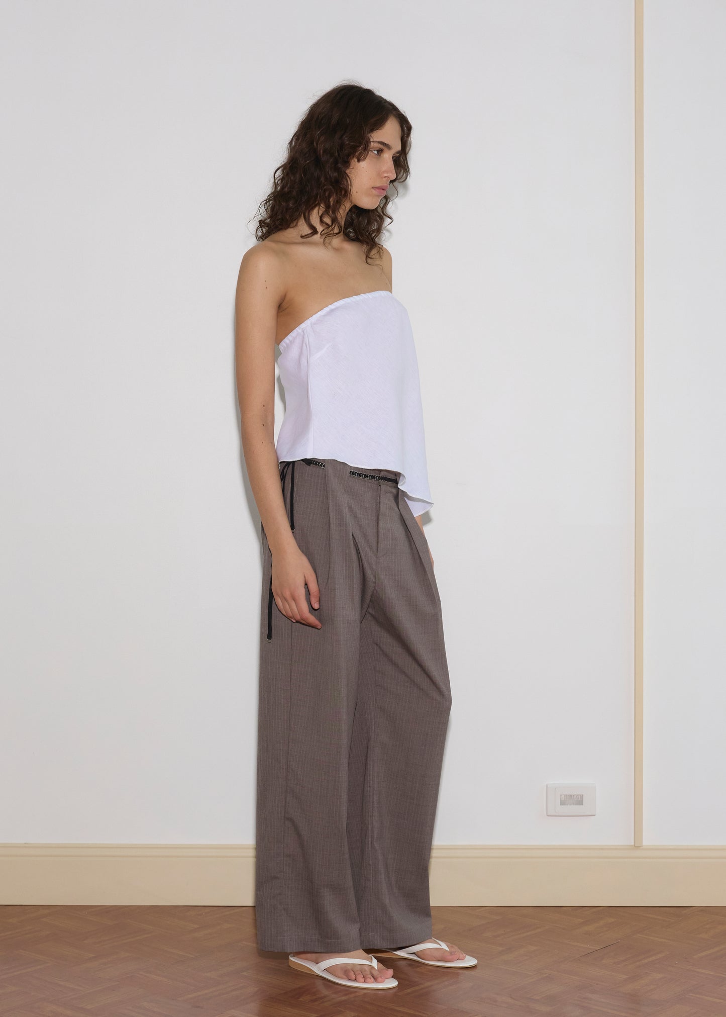 A person with curly brown hair stands indoors against a white wall, wearing a white strapless asymmetrical top, loose gray plaid trousers styled with a black Ring Loop Belt, and white sandals, maintaining a neutral expression.