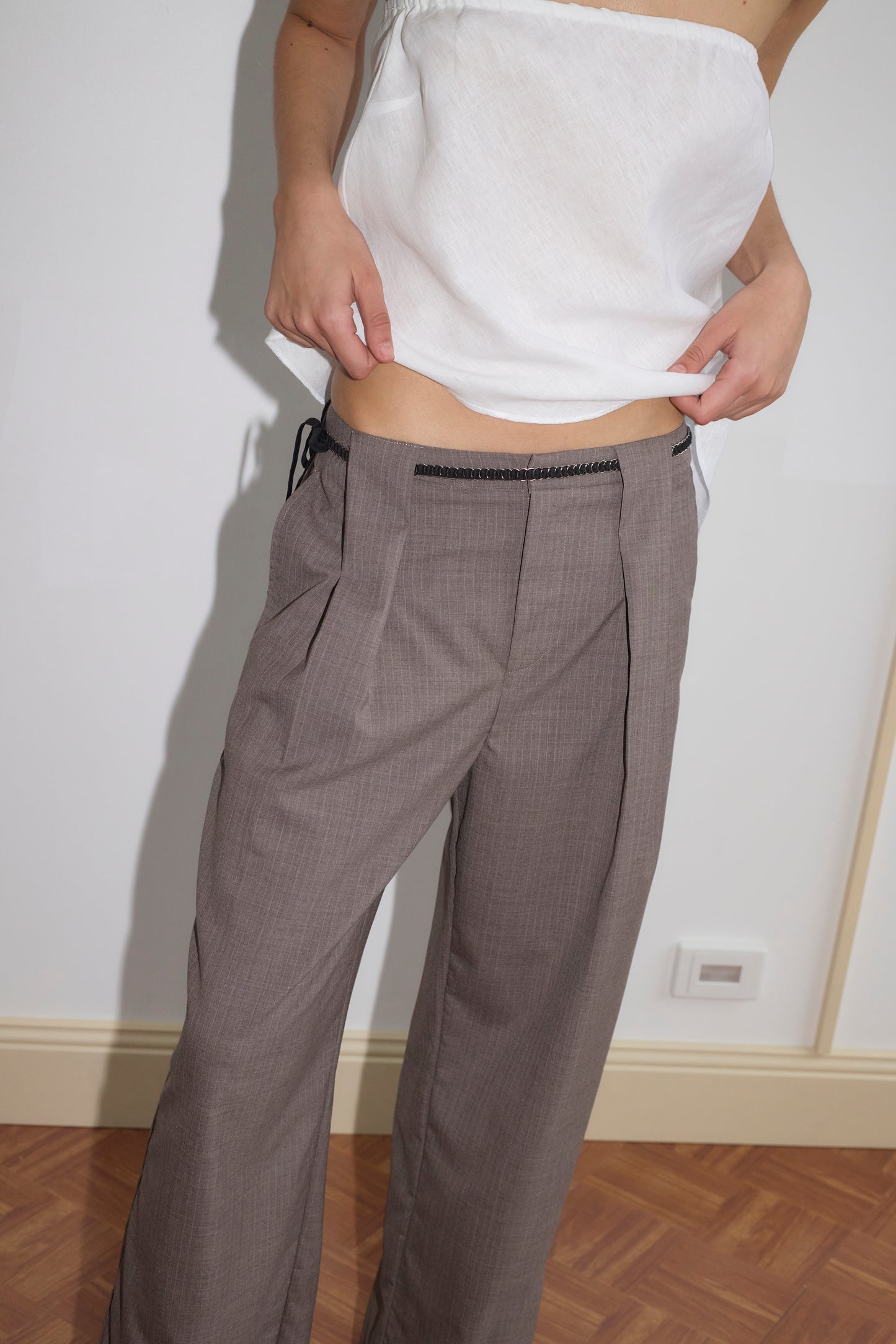A person wears a white strapless top and loose gray dress pants paired with the Ring Loop Belt - Black, standing indoors with hands in pockets and their midriff partially visible.