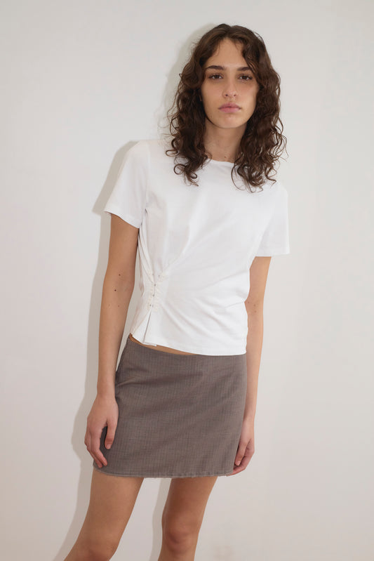 A young woman with wavy brown hair wears the Pinch Tee - White, a relaxed short sleeve top with a classic crew neckline and ruched details, paired with a gray mini skirt. She stands against a plain white background, looking at the camera.