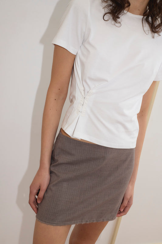 Wearing the Pinch Tee - White, a relaxed short sleeve top with a classic crew neckline and side button details, paired with a grey mini skirt. The person is standing against a plain light background, face not visible.