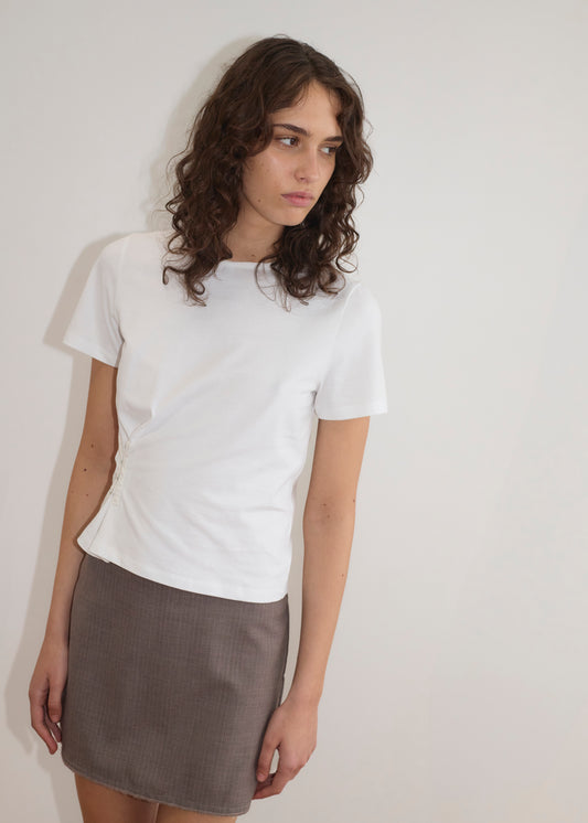 A person with wavy brown hair wears the Pinch Tee - White, featuring a classic crew neckline and relaxed short sleeves, paired with a gray skirt. They stand against a plain white wall, looking slightly to the side with a neutral expression.