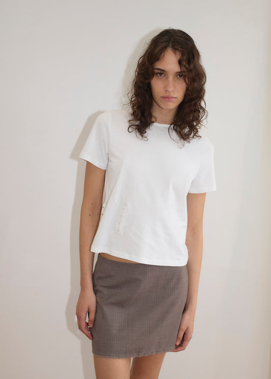 A young woman with wavy brown hair stands against a plain white wall, wearing the Pinch Tee - White with a classic crew neckline and a gray mini skirt. She looks slightly down at the camera, her expression neutral.
