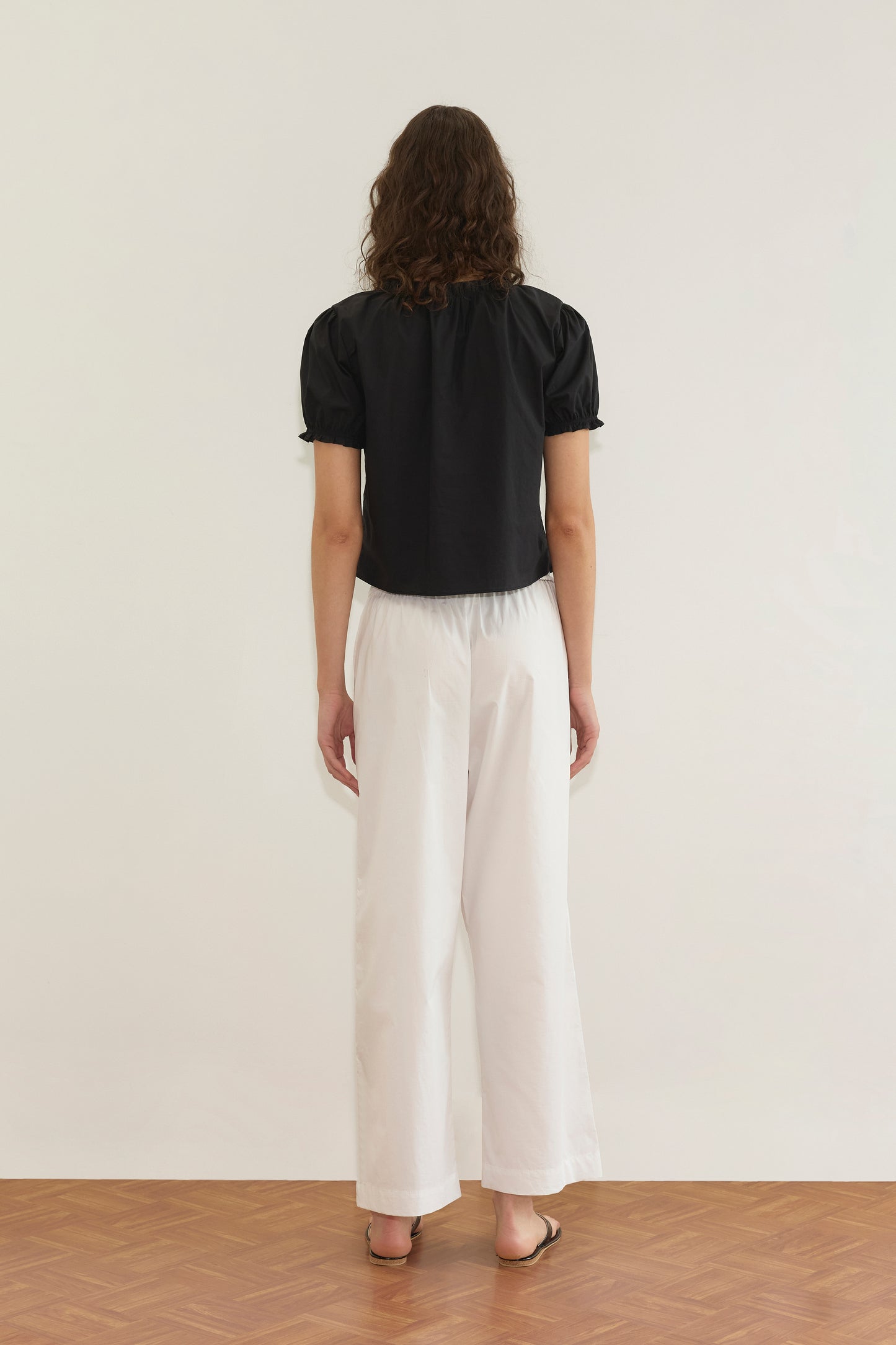 A person with wavy brown hair stands facing away, wearing the Cross Frill Top - Black with a cropped relaxed silhouette, paired with loose white pants and brown sandals on a wooden floor against a plain white wall.