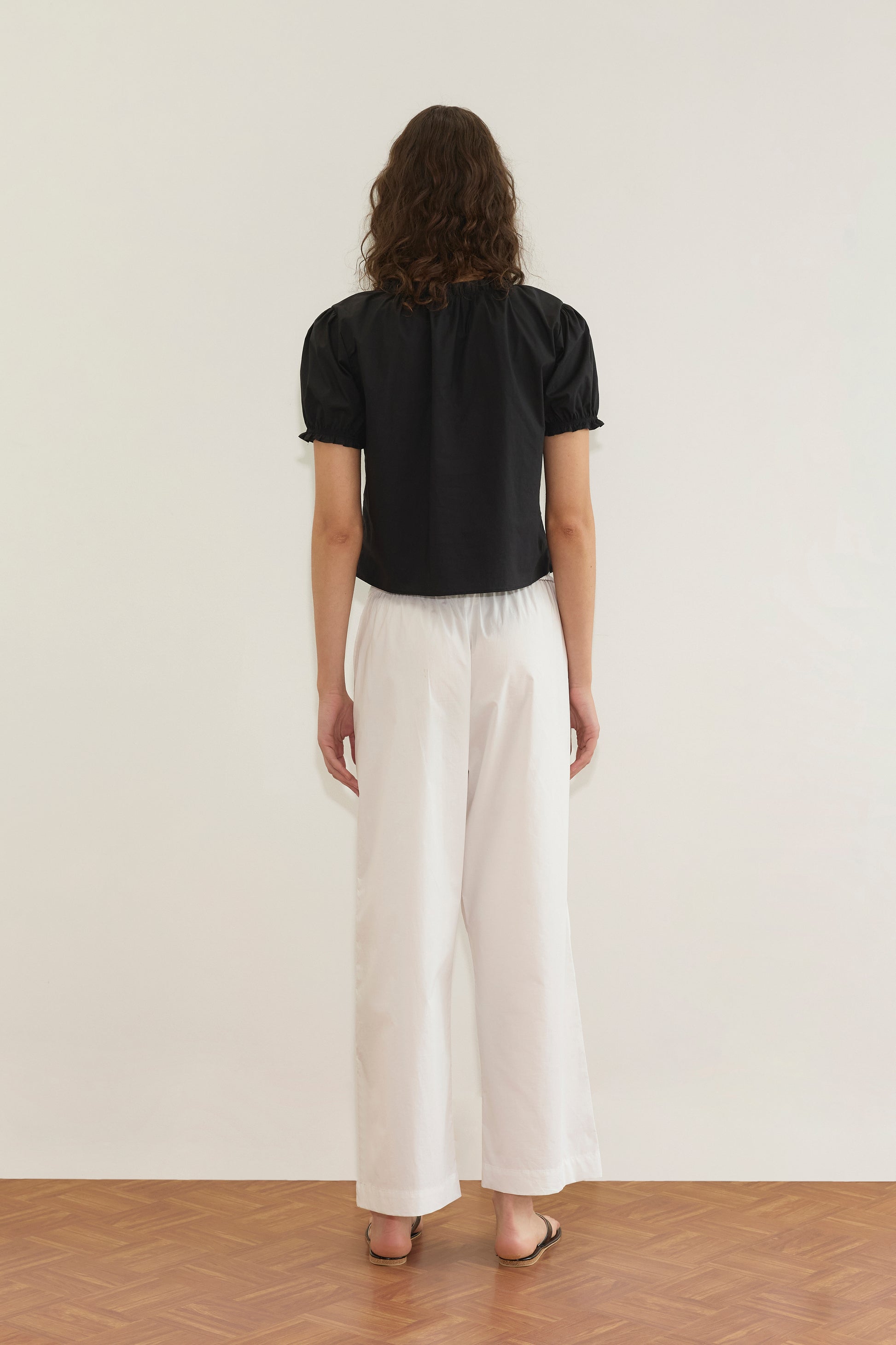 A person with wavy brown hair stands facing away, wearing the Cross Frill Top - Black with a cropped relaxed silhouette, paired with loose white pants and brown sandals on a wooden floor against a plain white wall.