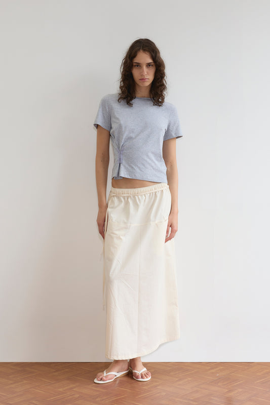 A person with long curly hair stands indoors against a white wall and wooden floor, wearing a Deiji Studios Pinch Tee - Grey, a cream long skirt with pockets, and white sandals.
